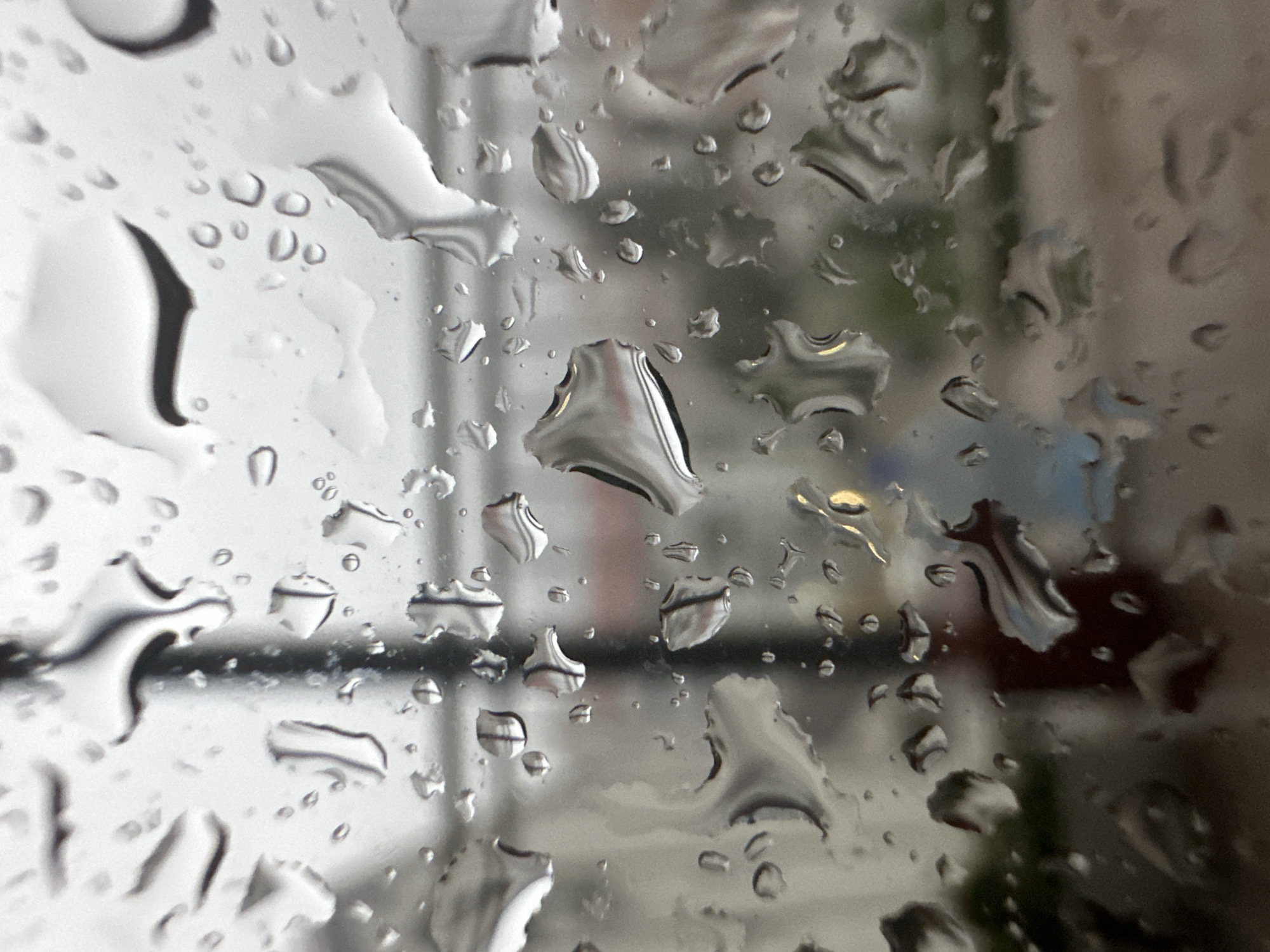 The image displays a close-up, highly detailed view of a transparent surface, likely glass, heavily covered with numerous individual raindrops of varying sizes and shapes. The raindrops are distinct and in sharp focus, reflecting and refracting ambient light, creating distorted visual effects. Through this rain-streaked foreground, a blurred, out-of-focus urban environment in Budva, Montenegro, is visible in the background. Distinguishable elements in the background include indistinct architectural forms, characterized by shades of grey and muted tones, suggesting buildings or structures. A prominent dark vertical element, possibly a pole or part of a railing, is visible towards the center-left. Faint horizontal lines further suggest distant building features. A small, distinct yellowish-orange light source is observed in the middle-right portion of the background. Scattered, blurred patches of dark green suggest the presence of distant vegetation. A reddish-brown area is also vaguely discernible in the upper-right quadrant. No individuals or specific human activities are visible within the scene. The overall impression is that of viewing an urban landscape during rainfall or overcast weather conditions.