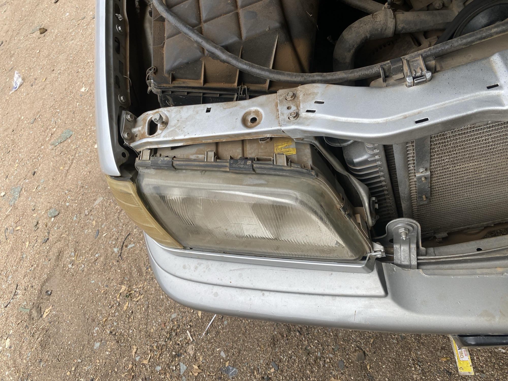 The image shows a close-up, overhead view of the front passenger side of a silver car, possibly a Mercedes-Benz, with the headlight assembly detached and lying on the ground. The car's front fender and part of the engine bay are visible. The ground is covered with dirt and small pebbles, suggesting an outdoor setting. The car appears to be in a state of disrepair or undergoing maintenance, with various mechanical components exposed. The scene is likely located in Jalingo, Nigeria, where such scenes are common in workshops or informal repair areas.