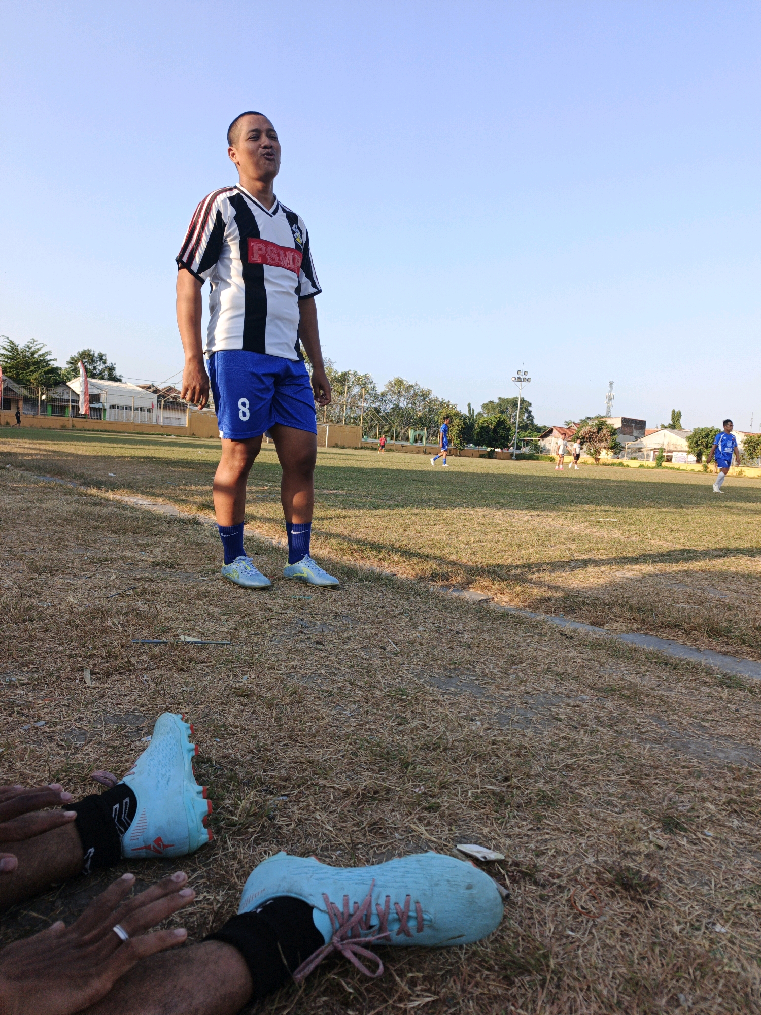 In Mojokerto, Indonesia, on a dry, grassy soccer field under a clear sky, a male soccer player stands facing forward, looking upwards with his mouth open. He wears a black, white, and red striped jersey with "PSMP" printed on it, blue shorts with the number "8" on the left leg, blue socks, and light blue soccer cleats. In the foreground, the feet of another person wearing similar light blue soccer cleats and black socks are visible, with one hand resting near the cleats. Other players in blue and white uniforms can be seen on the field in the background, and a building and trees are visible beyond the field.