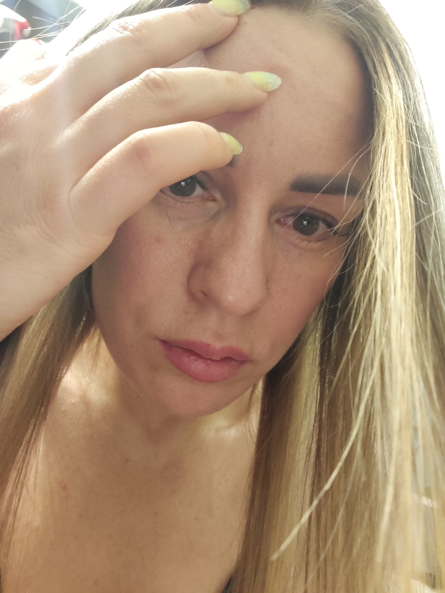 The image is a close-up, self-portrait style photograph of a woman's face and upper chest. She has long, blonde hair that is partially covering her face. Her right hand is raised and touching her forehead. She is wearing light-colored nail polish. Her eyes are open and looking towards the camera. Her lips are slightly parted and appear to be painted with a light pink lipstick. There are some small blemishes or moles visible on her skin. The lighting suggests an indoor environment, possibly with natural light coming from the left. There is no visible indication of the city of Almaty, Kazakhstan, or any specific urban environment.