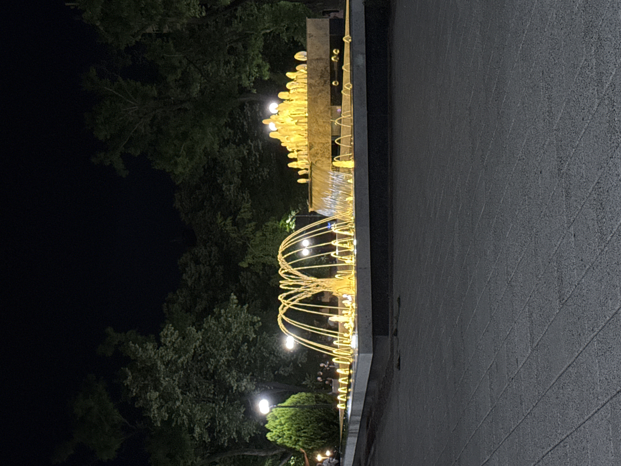 The image depicts a lit fountain at night in the city of Stara Zagora, Bulgaria. The fountain is illuminated with warm yellow lights, creating a visually striking effect. It features multiple water jets, some forming arched patterns and others cascading down. The fountain is situated within a rectangular pool with a stone border, and beyond that, a paved walkway. In the background, there are trees and streetlights, with a few people visible in the distance. The overall scene suggests a public park or square setting, with the fountain as a central point of attraction, likely a popular gathering spot in the evening.