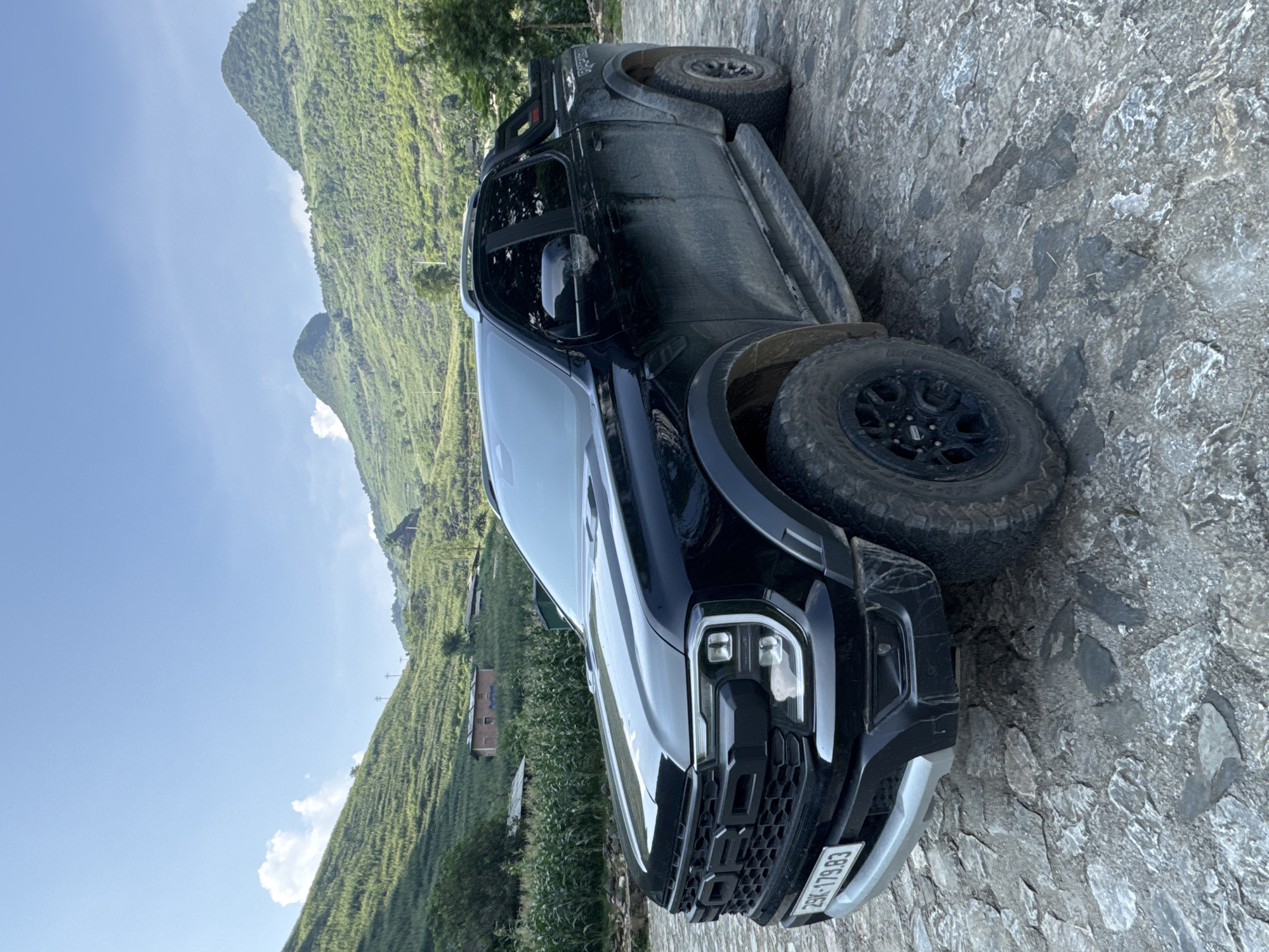 A black Ford Ranger pickup truck, covered in dirt, is parked on a stone road. The truck is oriented diagonally in the frame. The background features a mountainous, green landscape with terraced fields. A small building is also visible on the hillside. The photograph was taken in Đường Thượng Commune, Vietnam. The vehicle's license plate is visible. The lighting is bright and appears to be daylight.