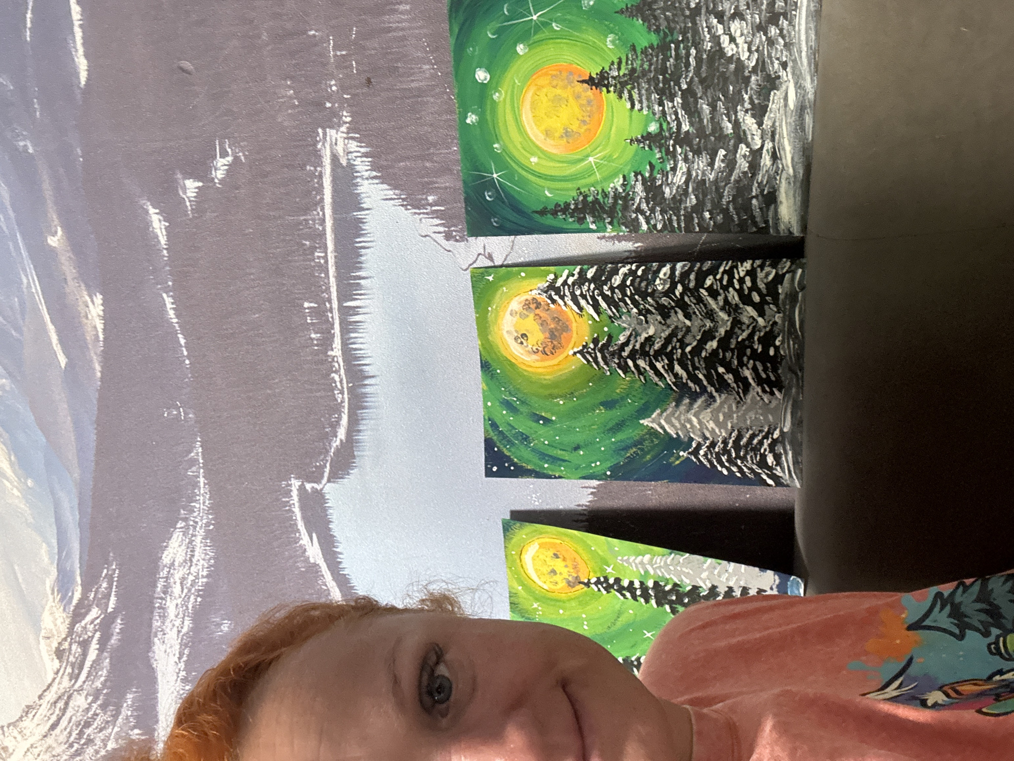 A woman with fair skin and red hair is taking a selfie in front of a wall. The wall has a mural of what appears to be snow-capped mountains, and three small paintings of a full moon and a coniferous tree are hanging on it. The woman is wearing a peach-colored t-shirt with a graphic design on it. The edge of a dark-colored couch or chair is visible on the right side of the frame. The location is indeterminate; there is no evidence to suggest this photo was taken in Chaikovsky, Russia.