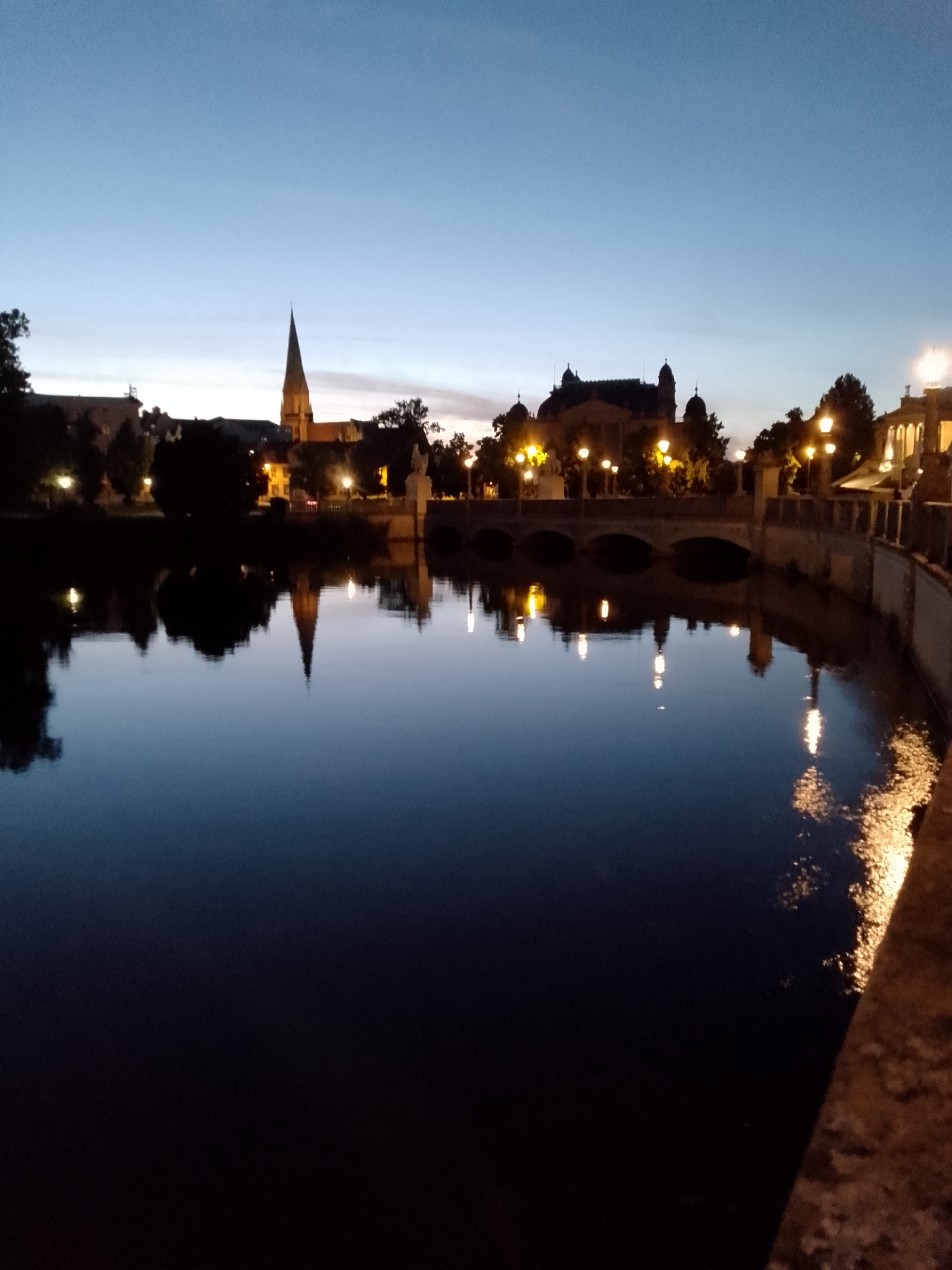 The image depicts a twilight scene over a body of water in Schwerin, Germany. The sky is a deep blue, transitioning to a lighter hue near the horizon, indicating either dawn or dusk. The water is dark and still, reflecting the lights and silhouettes of the buildings and structures on the opposite bank.
Prominent in the skyline is a tall, pointed spire, characteristic of a church or cathedral. To its right, larger buildings with domed roofs are visible, illuminated by a series of warm, yellow streetlights lining a bridge that spans the water. These lights create a beautiful reflection on the water's surface. The architecture suggests a historical European city.
The scene is devoid of people, contributing to a peaceful and tranquil atmosphere. The overall mood is serene and slightly melancholic, often associated with the transitionary hours of the day. The lighting suggests that the photograph was taken after sunset or before sunrise, with artificial lights beginning to dominate the natural light. The presence of a bridge and waterfront buildings strongly points to an urban setting.