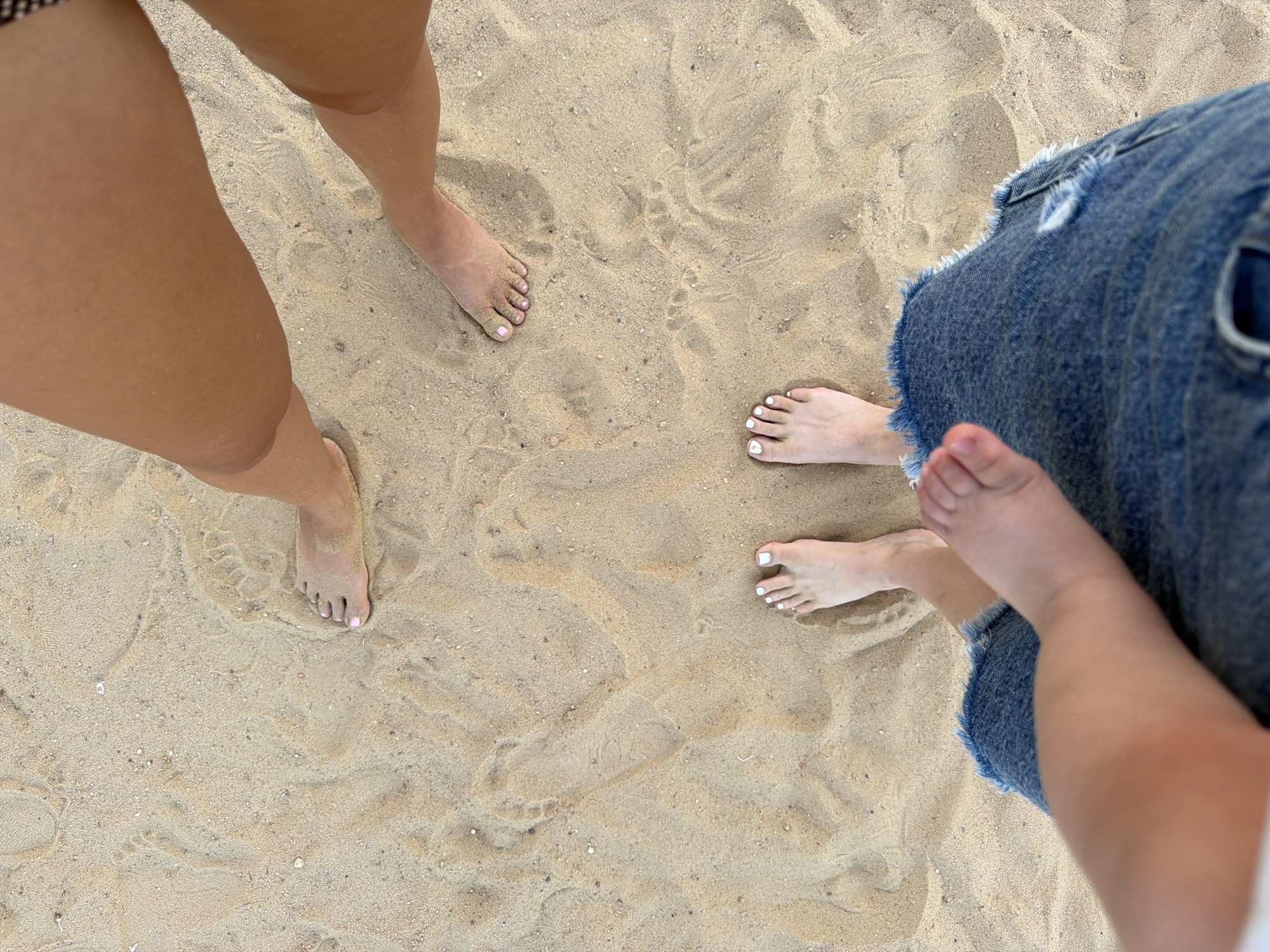 The image shows the lower legs and feet of at least three people on a sandy beach. The sand is light brown and appears dry, with subtle footprints and indentations visible.
On the left side of the frame, two bare feet and lower legs are visible. One foot has light pink nail polish on its toenails. The skin tone is fair.
On the right side of the frame, two more bare feet are visible, along with a portion of denim shorts or a skirt. One of these feet has white nail polish. The other foot is a smaller foot, likely belonging to a child, with no visible nail polish.
The scene suggests a casual outing to the beach, possibly on a sunny day given the lighting. The presence of multiple feet, including a child's, implies a group of people, perhaps a family, enjoying time together. The setting is clearly a beach with its characteristic sand.
There is no visible text in the image. The time of day and weather are not explicitly stated, but the lighting suggests it could be daytime. The location context provided is Sharm El Sheikh, Egypt, which aligns with a beach setting. The emotions conveyed are generally relaxed and summery due to the context of a beach outing.