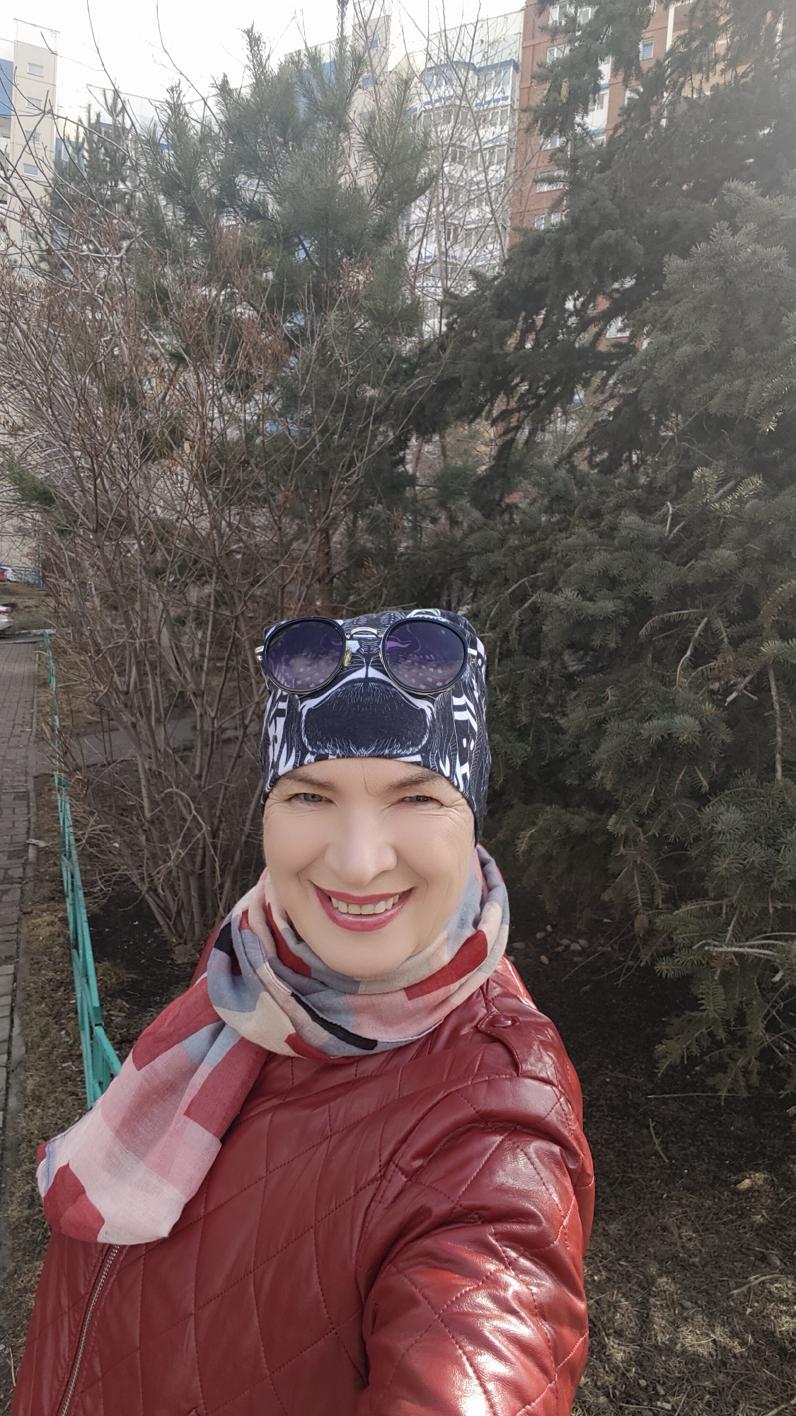 A woman is taking a selfie outdoors. She is wearing a patterned hat with sunglasses perched on top, a scarf with a geometric pattern, and a quilted red jacket. Her expression is a smile.
Behind her are two trees, one with bare branches and the other a green evergreen. A green metal fence and brick walkway are visible in the foreground on the left. In the background, a multi-story building is partially visible through the trees.
