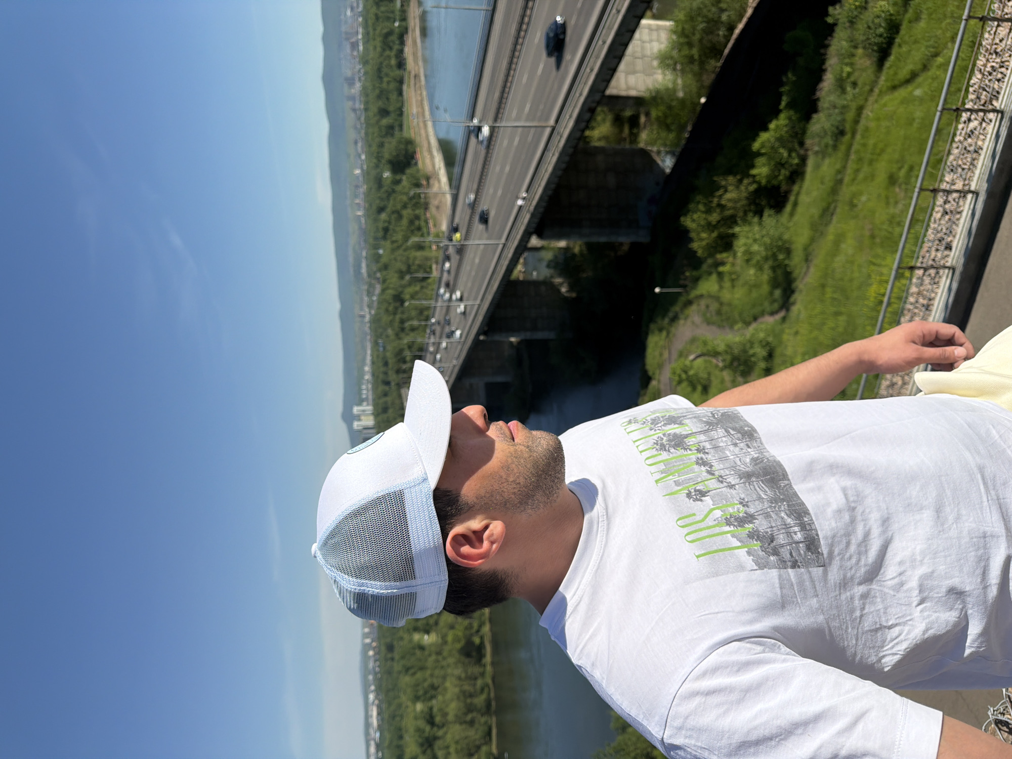 A male individual stands in profile on an elevated viewpoint, facing right from the viewer's perspective, overlooking the Yenisei River and the Nikolaevsky Bridge in Krasnoyarsk, Russia. He wears a white t-shirt featuring a graphic of "LOS ANGELES" in green text above a monochrome cityscape with palm trees, and a white baseball cap with light blue mesh side panels. His right arm is visible, hanging alongside his body.
The background shows a wide, dark blue river flowing beneath a multi-lane concrete highway bridge with multiple pillars. Vehicles are observable in motion on the bridge. Dense green forests and vegetation cover the riverbanks and the undulating terrain, extending to the horizon where urban structures of Krasnoyarsk are visible. The sky above is clear and bright blue with a few subtle, wispy cloud formations. A metal railing with a gravel base marks the edge of the elevated viewpoint in the lower right foreground. The scene is illuminated by strong sunlight, indicating a clear day.