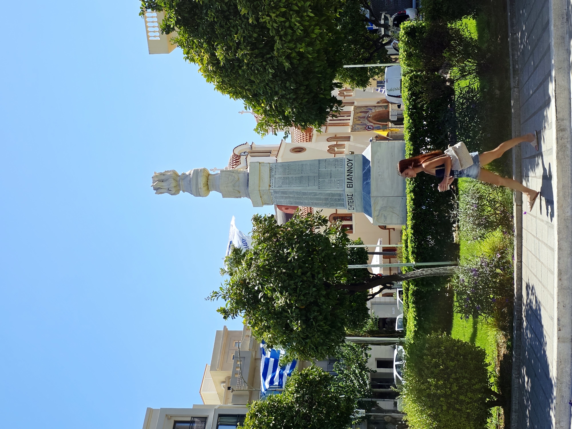 The image depicts a street scene in Agios Nikolaos, Crete, Greece, with a white marble monument as the central focus. The monument, which appears to be a memorial, features inscriptions in Greek. A young woman with long brown hair is walking past the monument from right to left, holding a cup in her right hand and carrying a light-colored woven bag over her left shoulder. She is wearing a dark blue tank top and denim shorts. Greek flags, one large blue and white striped flag and another white flag with blue lettering, are visible on flagpoles behind the monument and to the left, respectively. The monument is surrounded by green bushes and manicured grass. In the background, a cream-colored building with arched windows and a tiled roof, likely a church, is visible behind the monument. Modern buildings, trees, and parked cars are also present in the scene. The sky is clear and blue, indicating a sunny day. The ground in the foreground is paved with concrete tiles, with shadows cast by trees and possibly an overhead structure.