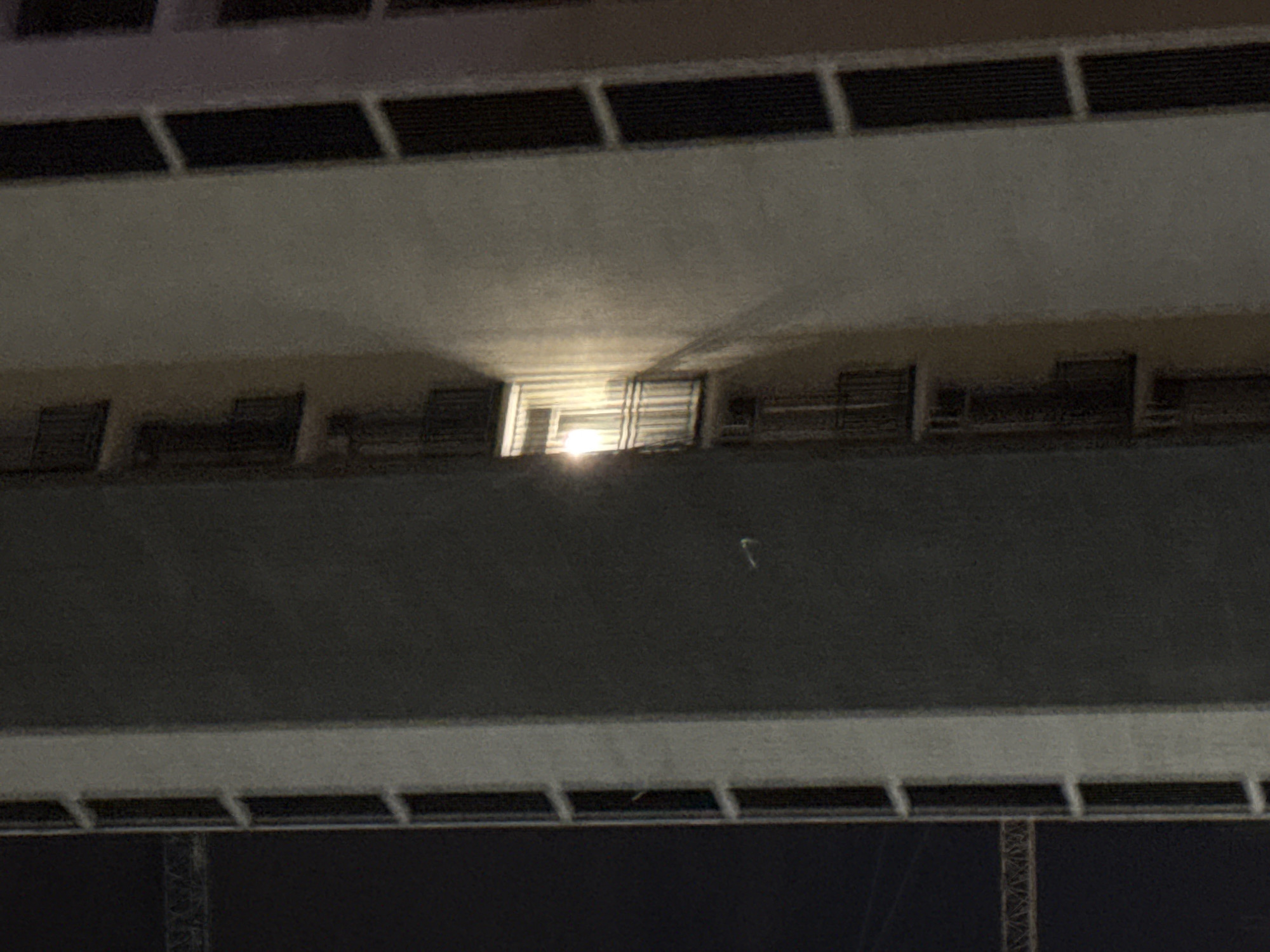 The image shows a close-up, night-time view of a modern apartment building in Tbilisi, Georgia. The building's facade is predominantly light-colored, with darker vertical elements that appear to be windows or balconies. One illuminated window is visible in the center of the frame, casting a bright light outwards. The building is tall, with multiple floors suggested by the repeating architectural elements. A construction crane is partially visible in the background to the left, indicating ongoing development or renovation in the area. The overall scene is urban and suggests a residential setting.