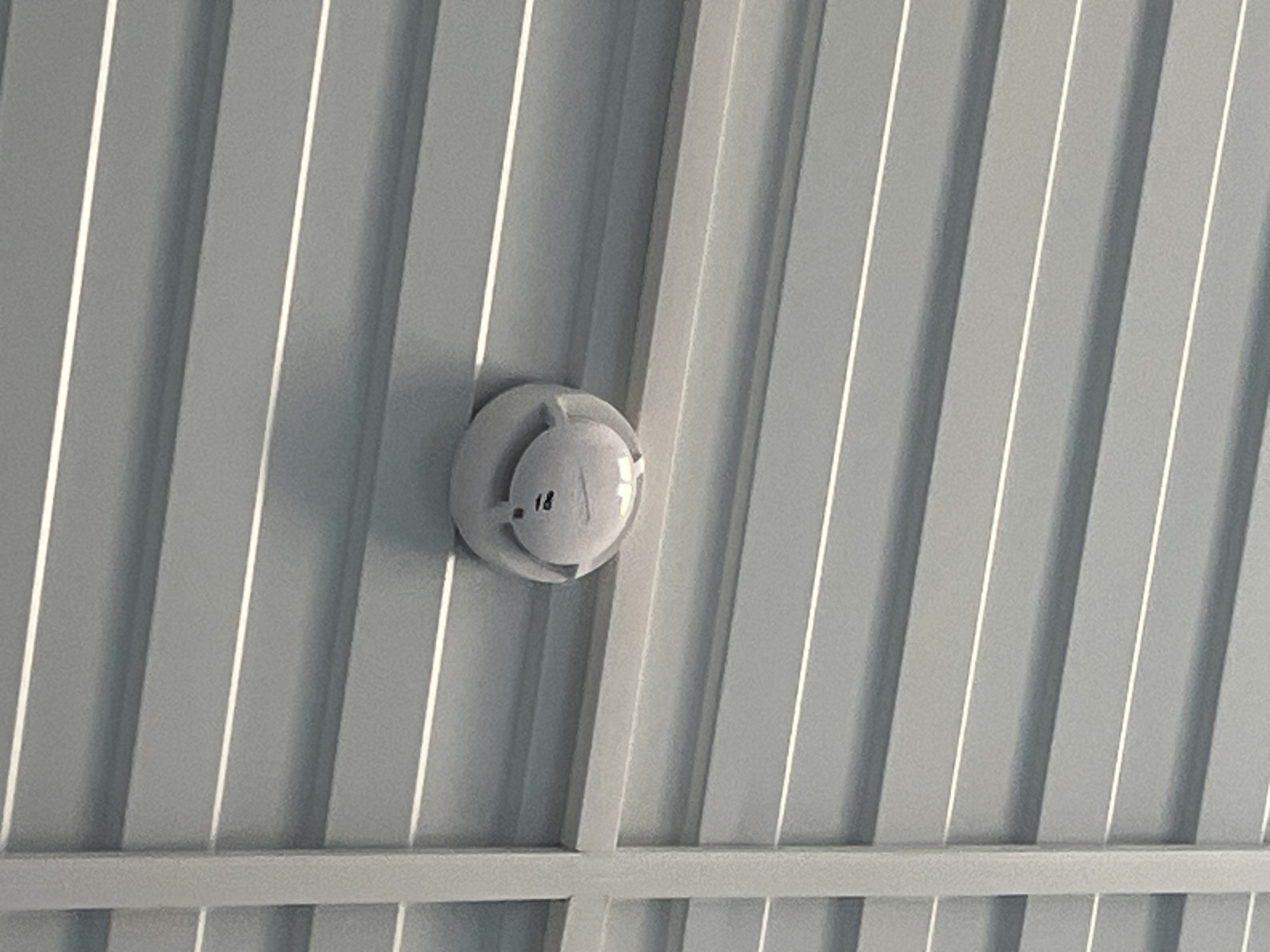 The image shows a white smoke detector mounted on a corrugated metal ceiling. The ceiling panels are grey with thin white lines along the corrugations. A vertical white metal beam is visible on the left side of the image, intersecting with the ceiling panels. The smoke detector has a slightly raised central section and appears to have some markings or buttons on its surface. The lighting suggests an indoor environment. There are no people or other objects of note in the image. The specific location in Usolye-Sibirskoye, Russia, is not determinable from the visual information provided.