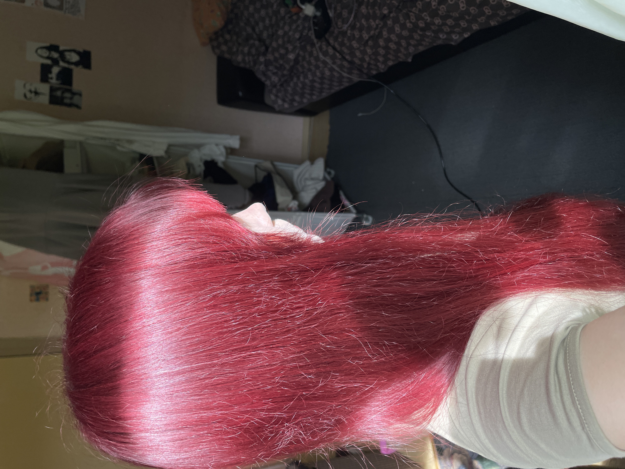 The image shows a close-up view of a person's bright red hair. The hair is long and appears to be styled with bangs that fall across the forehead. The person is wearing a light-colored, short-sleeved t-shirt. In the background, a bedroom is visible. There is a bed with patterned bedding, a dark-colored rug on the floor, and a closet or wardrobe with clothes hanging inside. On the wall, several black and white photographs are pinned up. The lighting suggests it is daytime, with sunlight casting a shadow on the person's shoulder and highlighting the hair. The scene is indoors, likely in a residential setting. The location of Солнечная Поляна, Russia is not evident from the image itself.
