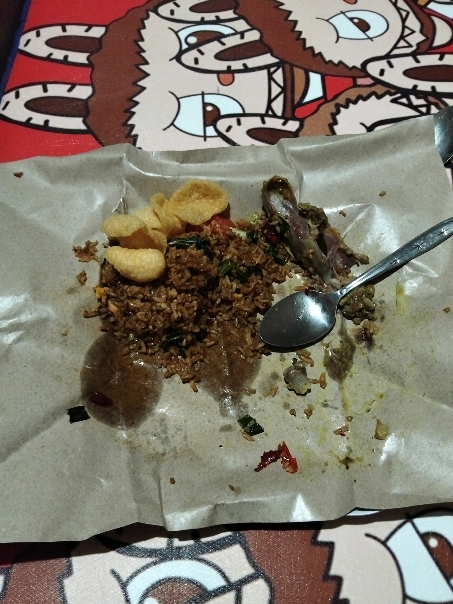 The image shows a close-up overhead view of a meal served on brown paper, possibly on a table with a cartoonish pattern in the background. The meal appears to be fried rice with a piece of chicken bone and some crackers or crisps. A spoon rests on the paper, suggesting the meal is partially eaten. The lighting suggests an indoor setting. There are no people visible in the image. The location within Panimbang, Indonesia, cannot be determined from the visual information provided.