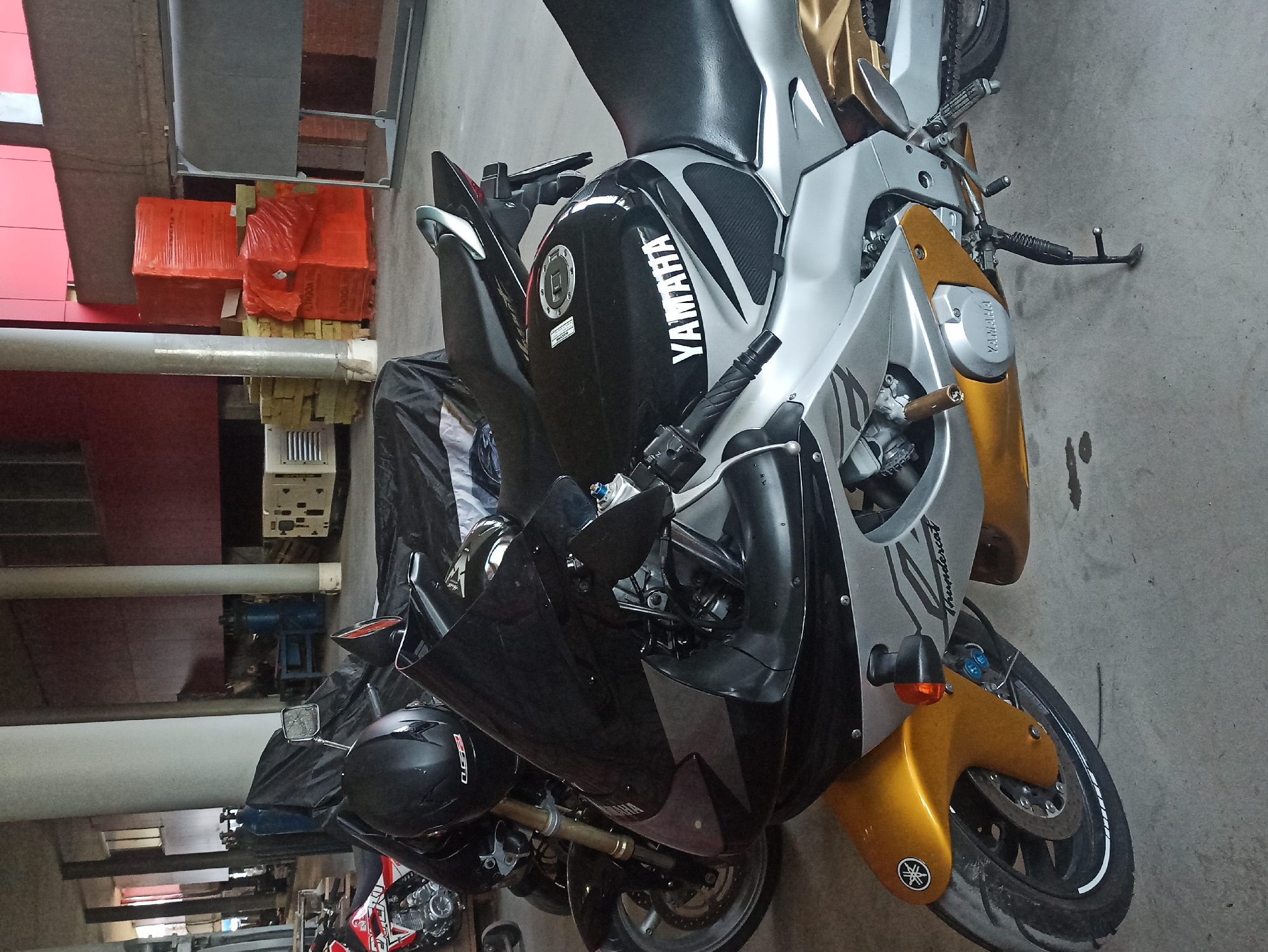 This image shows two Yamaha motorcycles parked in a garage. The main motorcycle is a black, silver, and gold Yamaha Thundercat with the Yamaha logo clearly visible on the fuel tank and fairings. A black helmet rests on the handlebars. The second motorcycle, only partially visible, is parked behind and to the left of the first. The environment appears to be a garage or storage area with various objects like orange wrapped items, machinery, and structural supports visible in the background. The location is unknown.