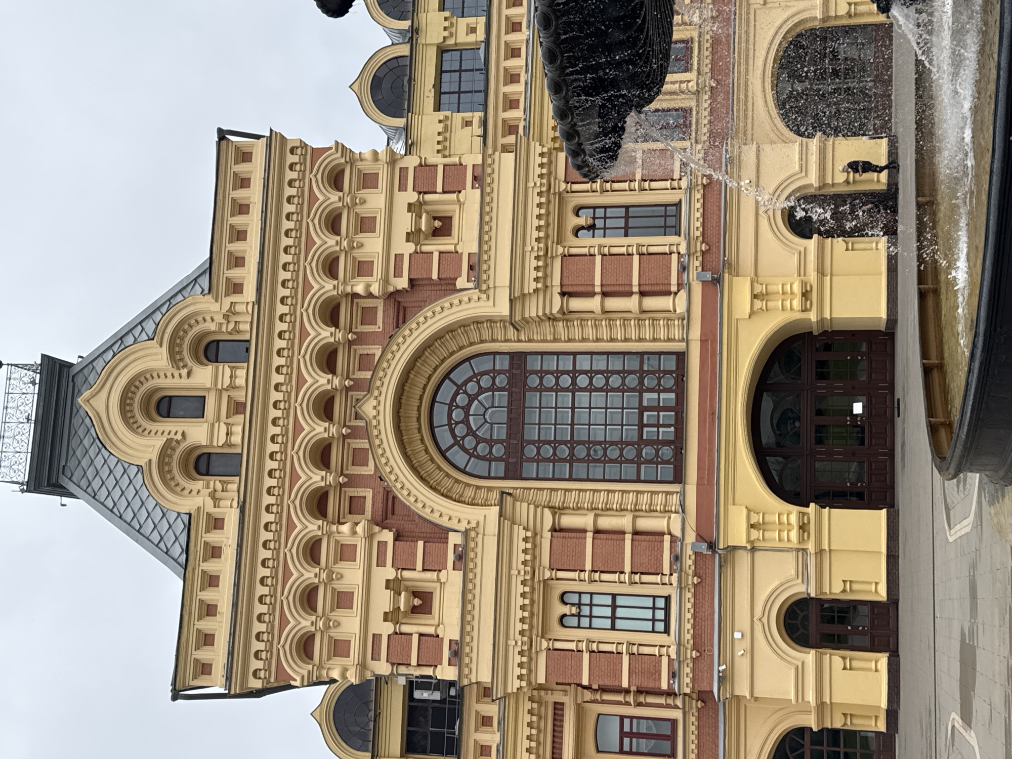The image depicts the Nizhny Novgorod Fair, a historic landmark in Nizhny Novgorod, Russia. The grand building, characterized by its vibrant yellow and red brick facade, showcases intricate architectural details. A prominent arched window with a dark wooden frame dominates the central portion of the building. Below, multiple arched doorways with wooden doors are visible, with one of them leading into the building.
In the foreground, a fountain with dark, ornate stonework is active, with water cascading from it. A lone figure, dressed in dark clothing, stands near an arched doorway to the right of the fountain. The ground in front of the building is paved with grey stone tiles. The sky is overcast, suggesting a cloudy day. The overall scene conveys a sense of historical grandeur and public space.