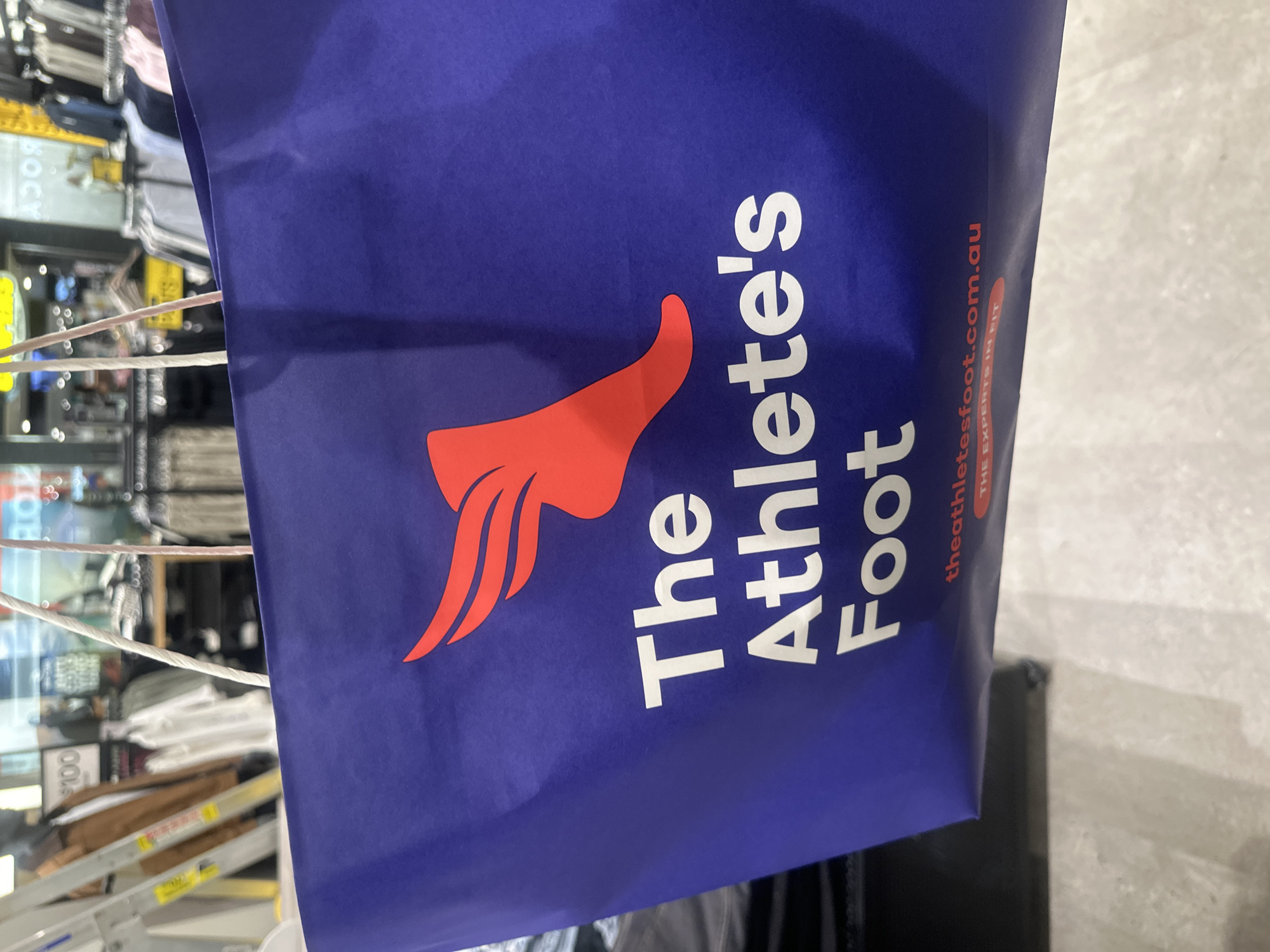The image shows a close-up of a blue shopping bag from "The Athlete's Foot". The bag features a red winged foot logo and the text "The Athlete's Foot" in white lettering. Below this, it says "theathletesfoot.com.au" and "THE EXPERTS IN FIT" in red. The background is blurred and appears to be inside a retail store, with racks of clothing and a price tag visible. The lighting suggests an indoor environment, likely during the day. This image was captured at the Big W store located in the Park Beach Plaza shopping center in Coffs Harbour, Australia.