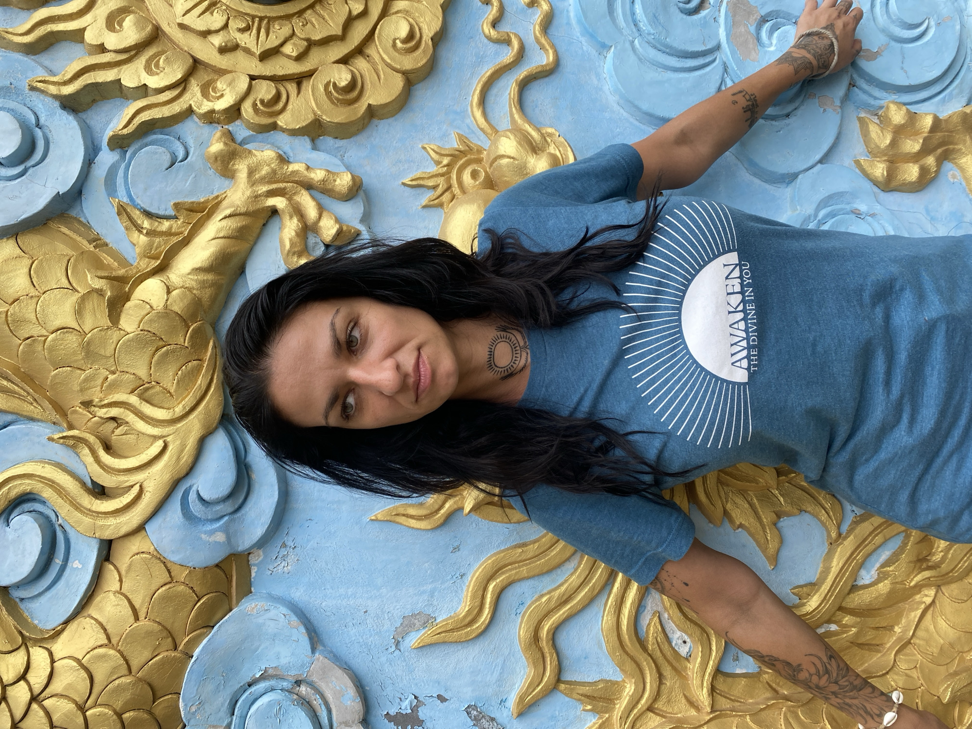 A woman with dark hair lies against a decorated wall in Mũi Né, Vietnam. The wall is adorned with gold and blue sculptural elements resembling a stylized dragon and clouds. She wears a blue t-shirt with the words "AWAKEN THE DIVINE IN YOU" printed on it. Tattoos are visible on both arms and her neck. Her expression is neutral as she looks toward the viewer.