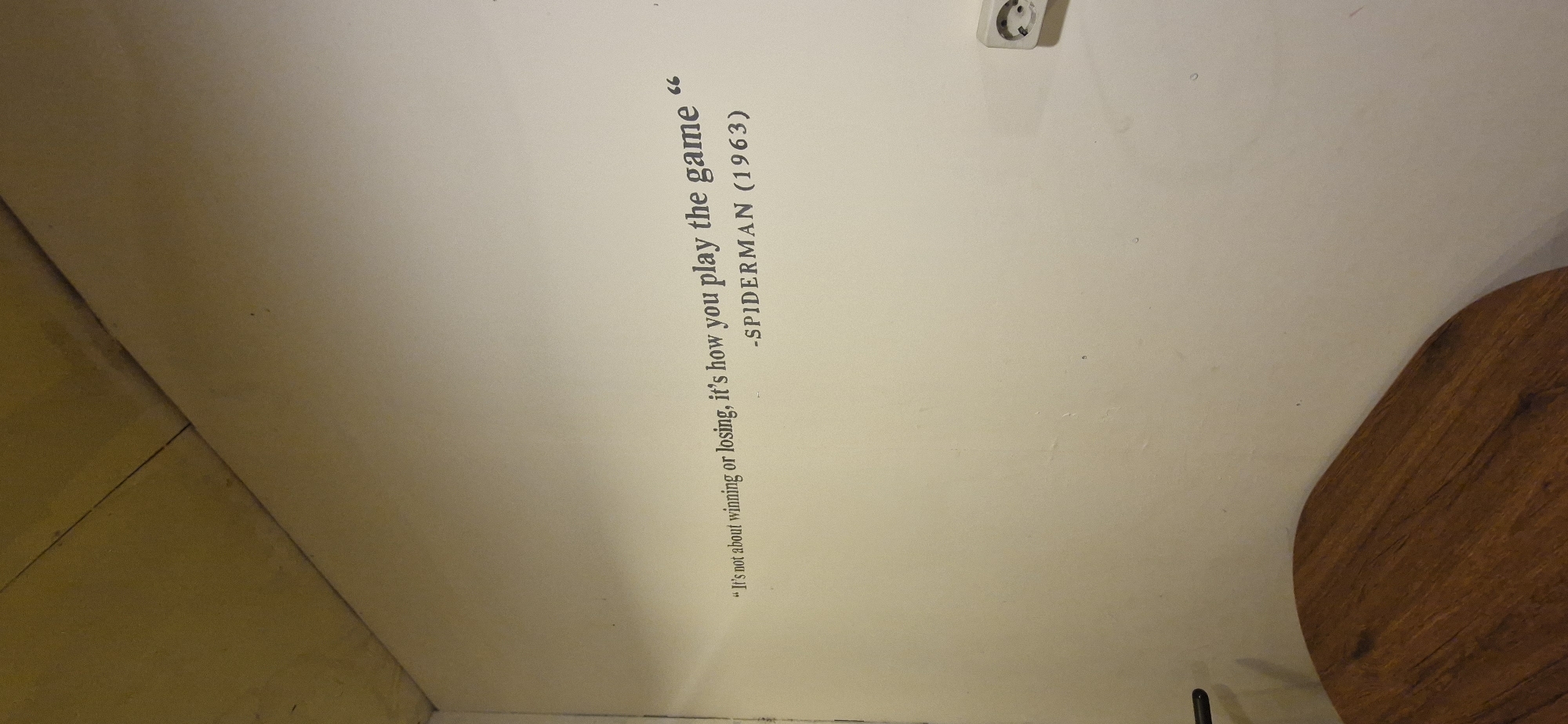 The image shows a wall in what appears to be an indoor setting, possibly a room or a cafe. A quote is stenciled onto the wall in dark gray text. The quote reads: "It's not about winning or losing, it's how you play the game" followed by "-SPIDERMAN (1963)". The wall is painted a light beige color and shows some minor imperfections. Above the wall, a section of a yellow ceiling or soffit with visible seams is present. In the lower left corner, the edge of a round, wooden table is visible. To the right of the quote, a white electrical outlet with a circular cover is mounted on the wall. The lighting suggests an indoor environment, and there are no people visible in the frame. The context of Rantau, Indonesia, is not visually supported by the image content.