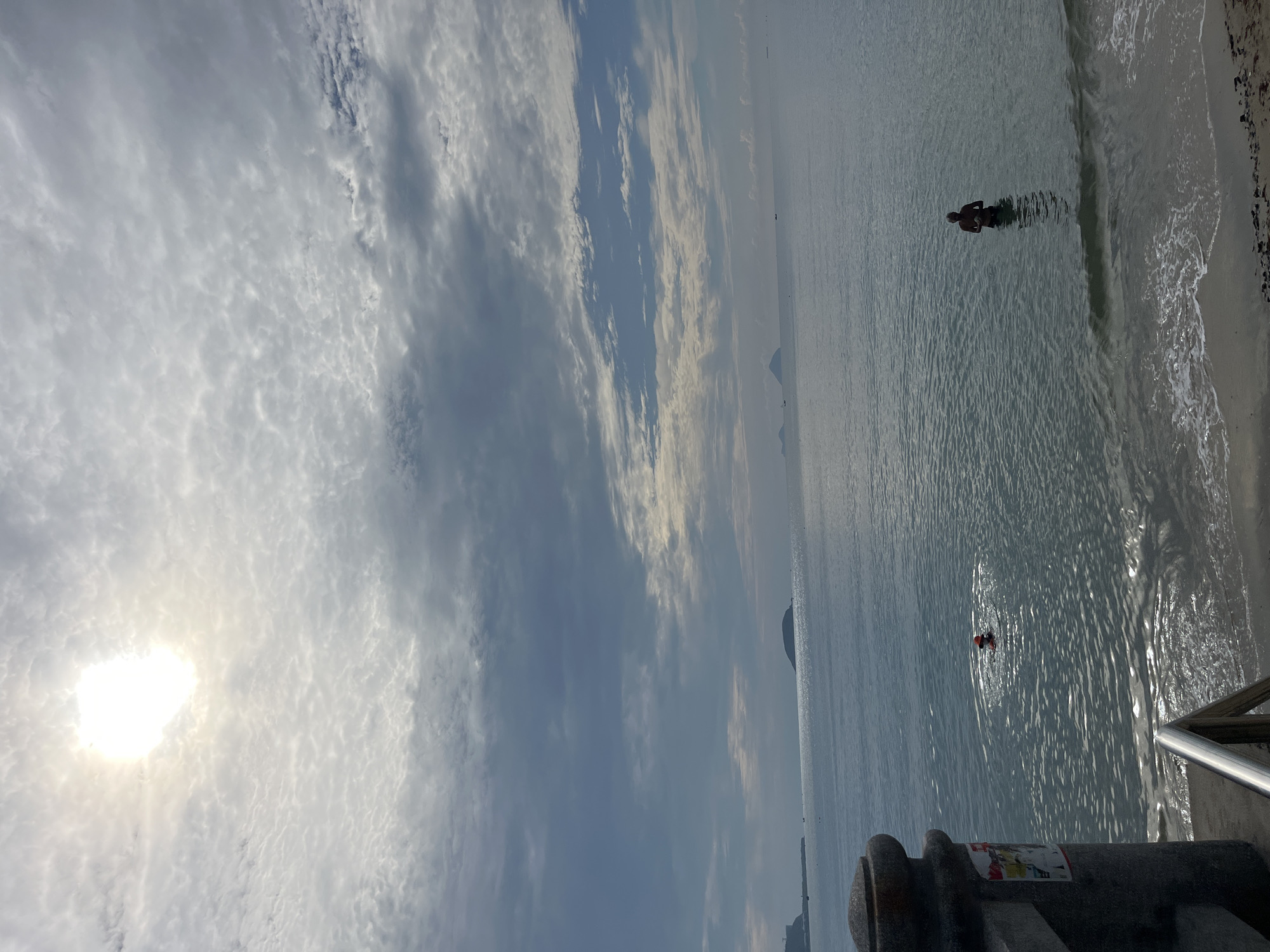 The image shows a beach scene with a cloudy sky above the ocean, taken in Bac Nha Trang Ward, Vietnam. The beach is visible on the right side of the frame, and a person is standing in the water near the shoreline. Further out in the water, another person is visible wearing an orange swim cap. The perspective is slightly tilted, and a concrete structure with a metal railing is situated in the lower right corner of the image.