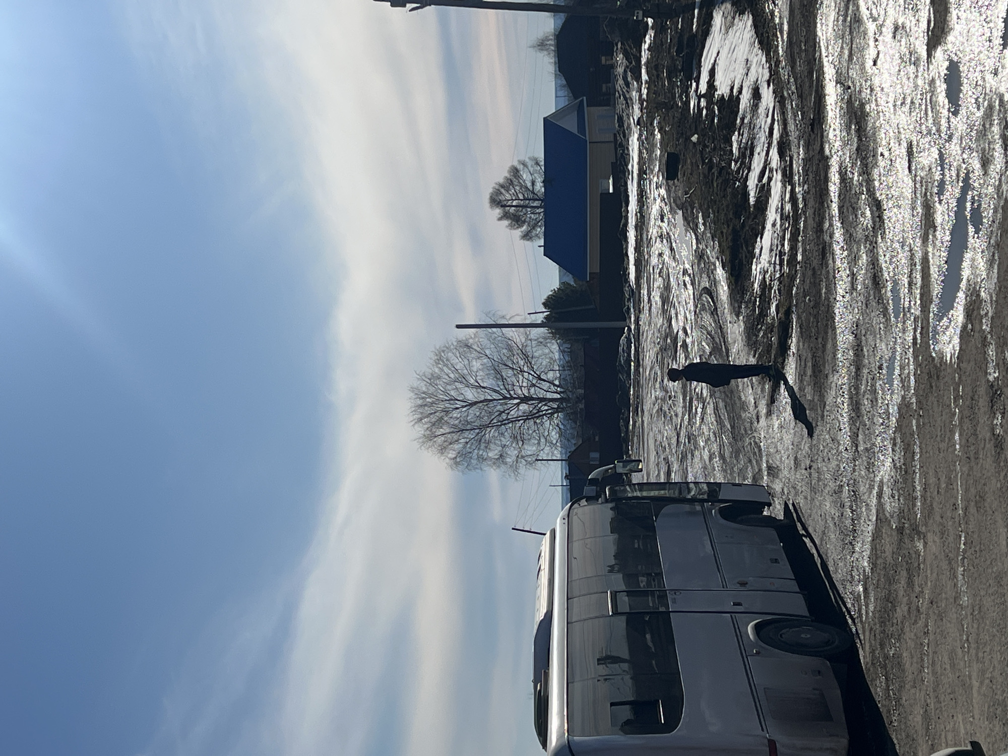 A horizontal, outdoor shot shows a partial view of a bus parked on a street next to a person walking on what appears to be a dirt or snow-covered road. The sky is light blue with light white clouds. In the background is a small house with a blue roof and bare trees. The road or pathway has some reflective patches, possibly from ice or water. The location is potentially Gory, Russia.