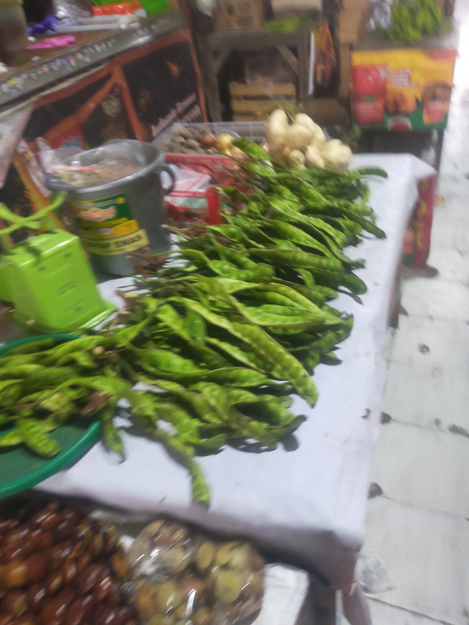 This is a forensic, matter-of-fact description of an image taken at a market stall in Trayeman, Indonesia.
The image depicts a market stall with various produce and items laid out on tables. In the foreground, a pile of green, elongated pods, likely stink beans (petai), is prominently displayed. To the left of the stink beans is a bright green mechanical scale. Behind the scale, a large grey bucket with a green label is visible. Further back, on a separate table, are some root vegetables, possibly radishes or jicama, and a basket of what appear to be onions. The background shows shelves with more items, including what appear to be packaged food products with colorful labels, and some hanging items. The overall environment suggests a busy market setting with goods for sale. No people are clearly visible or identifiable in the frame, and no specific actions or interactions are captured. The lighting is somewhat dim, consistent with an indoor market or an area with limited direct sunlight. The floor is a light-colored tiled surface.