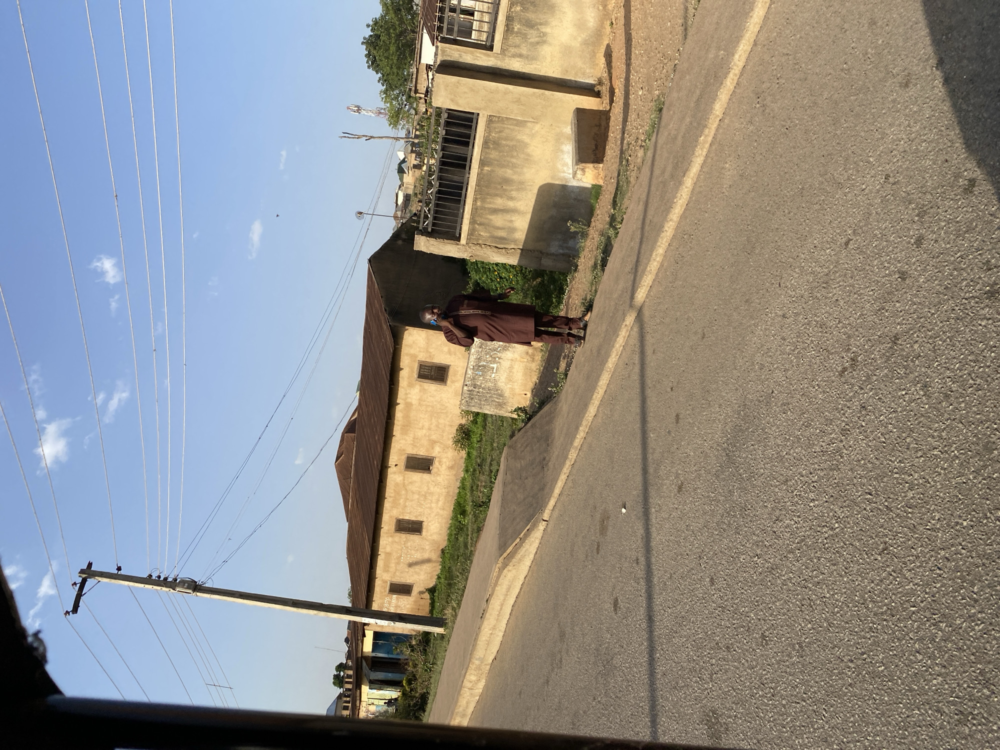 The image shows a street in Bukuru, Nigeria. A man wearing a brown robe and face mask stands on the sidewalk in front of a beige building with a rusty roof. A shorter concrete structure with a sign on it is to his left. Power lines are visible stretching into the sky. The street is asphalt with a concrete curb, and a shadow is cast on the right side of the frame. A utility pole is on the left side of the frame. Vegetation grows between the building and the sidewalk.