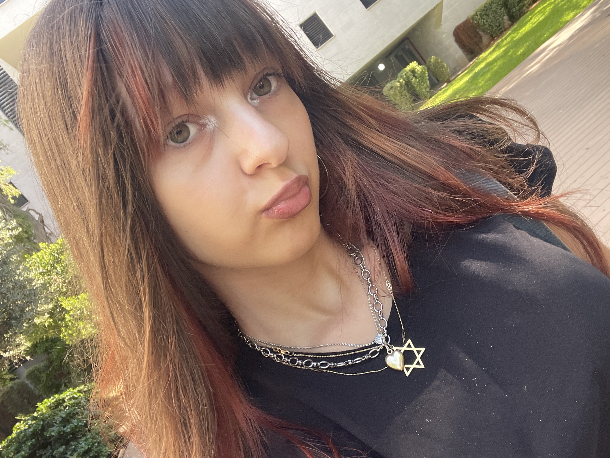 A young woman with long, brown hair with reddish highlights and bangs, is taking a selfie outdoors. She is wearing a black t-shirt and a layered necklace with a star of David pendant. Her lips are slightly pursed, and her eyes are open, looking towards the camera. The background shows greenery, a building with windows, and a paved walkway. The image appears to have been taken in the city of Ra'anana, Israel.