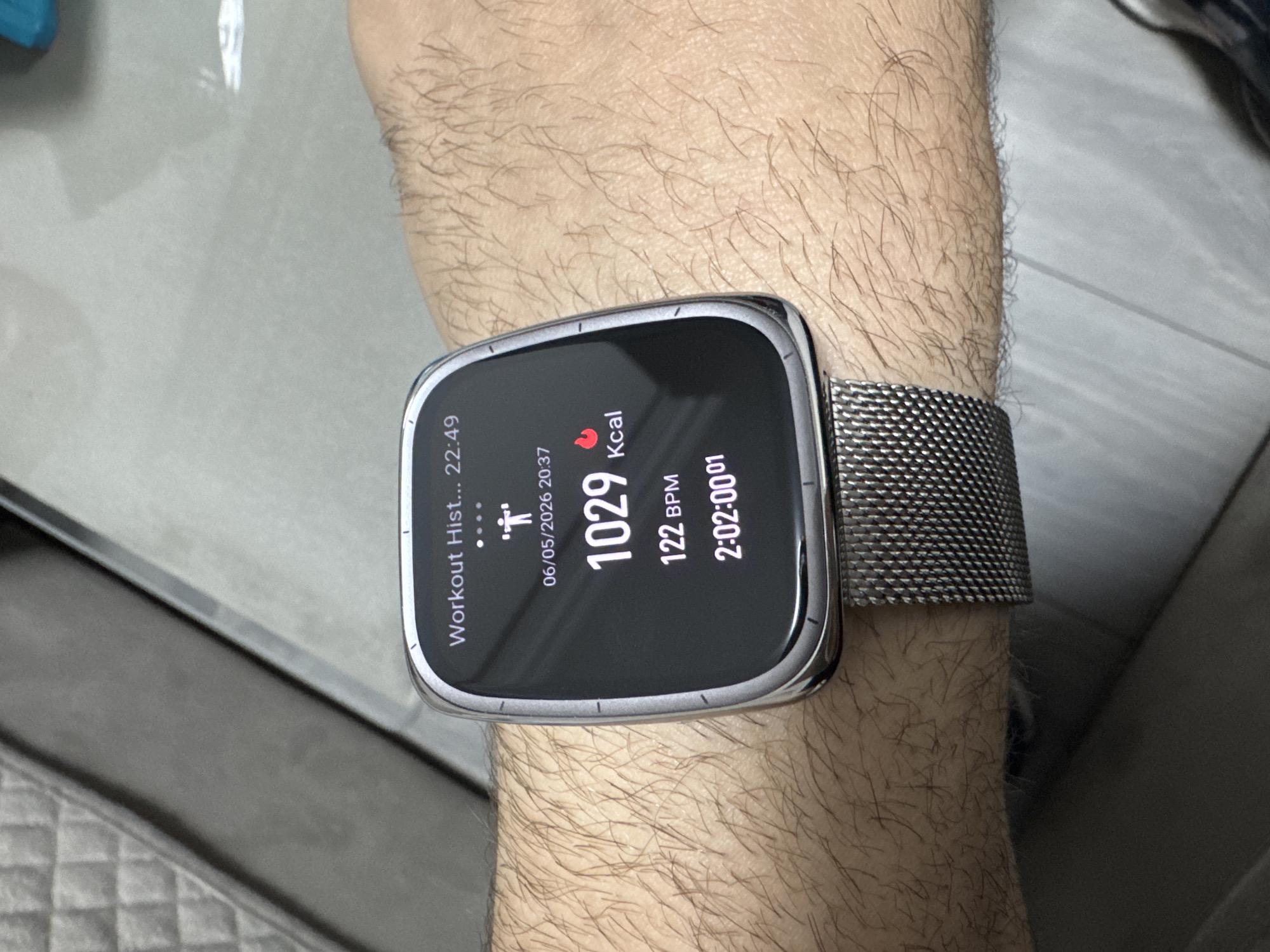 A close-up image shows a person's hairy wrist wearing a smartwatch. The smartwatch screen displays workout data: "Workout Hist... 22:49," a date "06/05/2026 20:37," "1029 Kcal," "122 BPM," and "2:02:0001." The watch has a silver metal mesh strap. The background is blurred and partially visible. There are no indications of the location being Yerevan, Armenia.