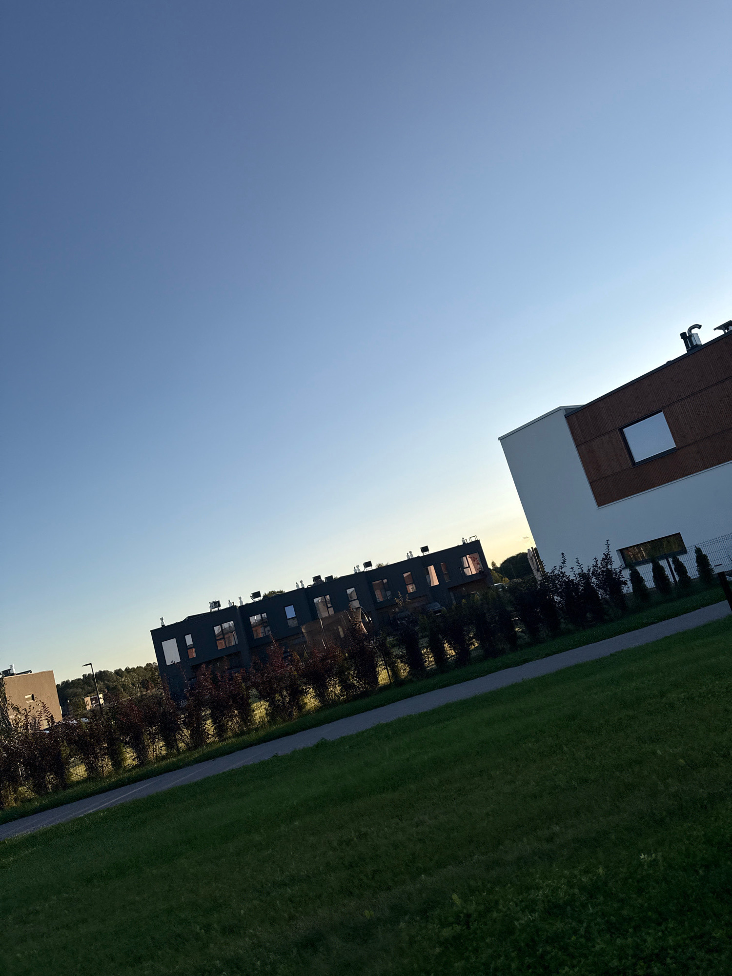 The image shows a view of modern residential buildings in Järveküla, Estonia, under a clear blue sky. In the foreground, a well-maintained green lawn stretches towards the bottom right corner of the frame, bordered by a paved pathway. Beyond the pathway, a row of dark green, dense bushes runs horizontally across the middle ground. Behind the bushes, a series of dark-colored, multi-story residential buildings are visible. These buildings feature numerous windows, some reflecting the sky. To the right, a contrasting building with white and brown facade elements and a prominent rectangular window is also present. In the background, more trees and the outline of other structures can be seen against the horizon. The lighting suggests it is either early morning or late afternoon, with a soft glow on the buildings. There are no people or animals visible in the scene.