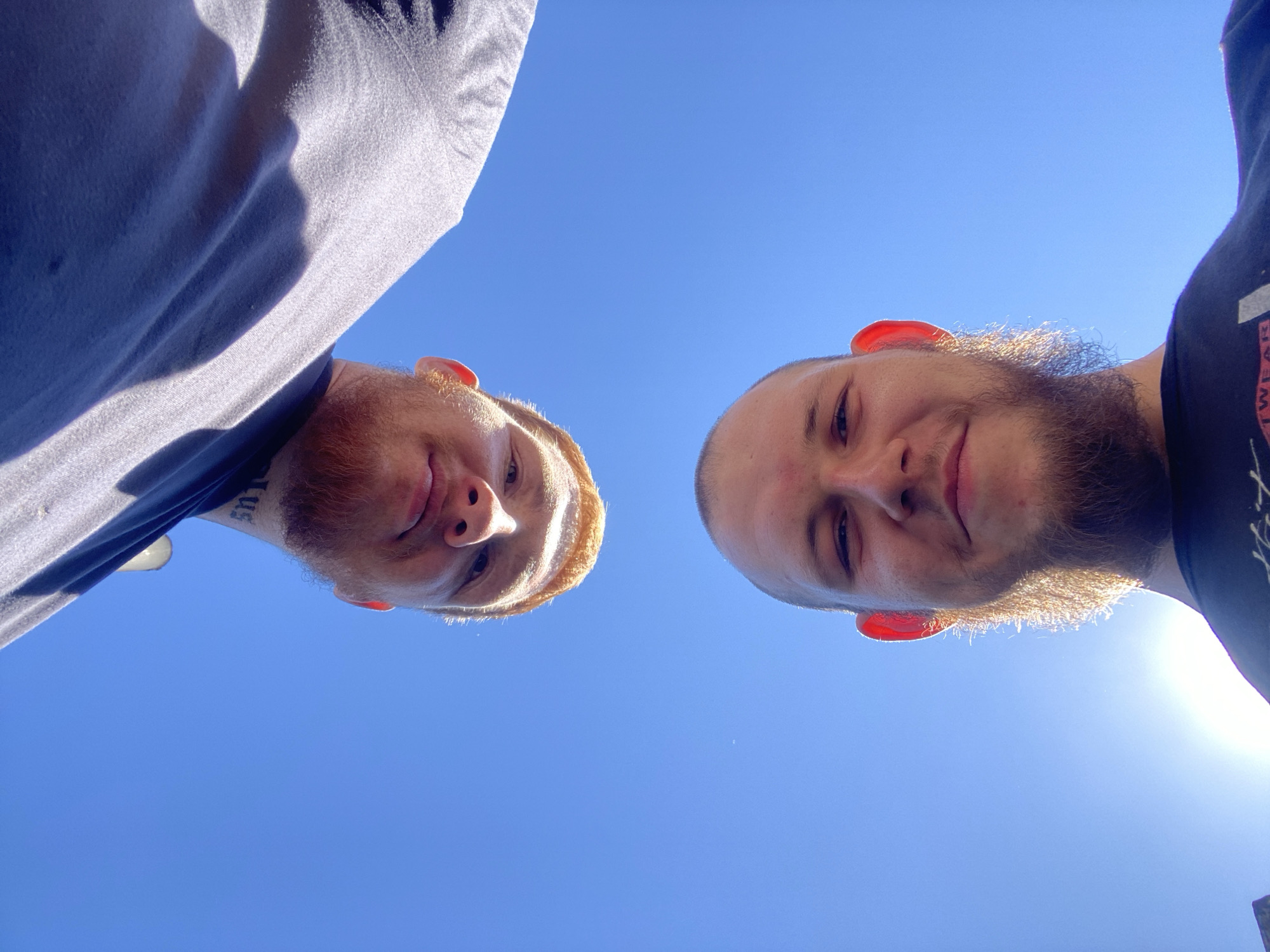 A low-angle, extreme wide shot shows two young adult Caucasian males against a clear blue sky, their faces positioned near the top and bottom of the frame respectively. Both men are looking up and directly at the camera. The male at the top is upside down, with his ginger beard and the word "LUS" tattooed on his neck visible. He has a slight smile. The male at the bottom is right-side up, also smiling. He has a short beard and a shaved head, with his ears appearing reddish, possibly from the sun. Both men are wearing dark t-shirts. The lighting suggests it is daytime, with the sun possibly behind the lower male, casting a bright glare. The background is entirely the sky, with no discernible city landmarks.