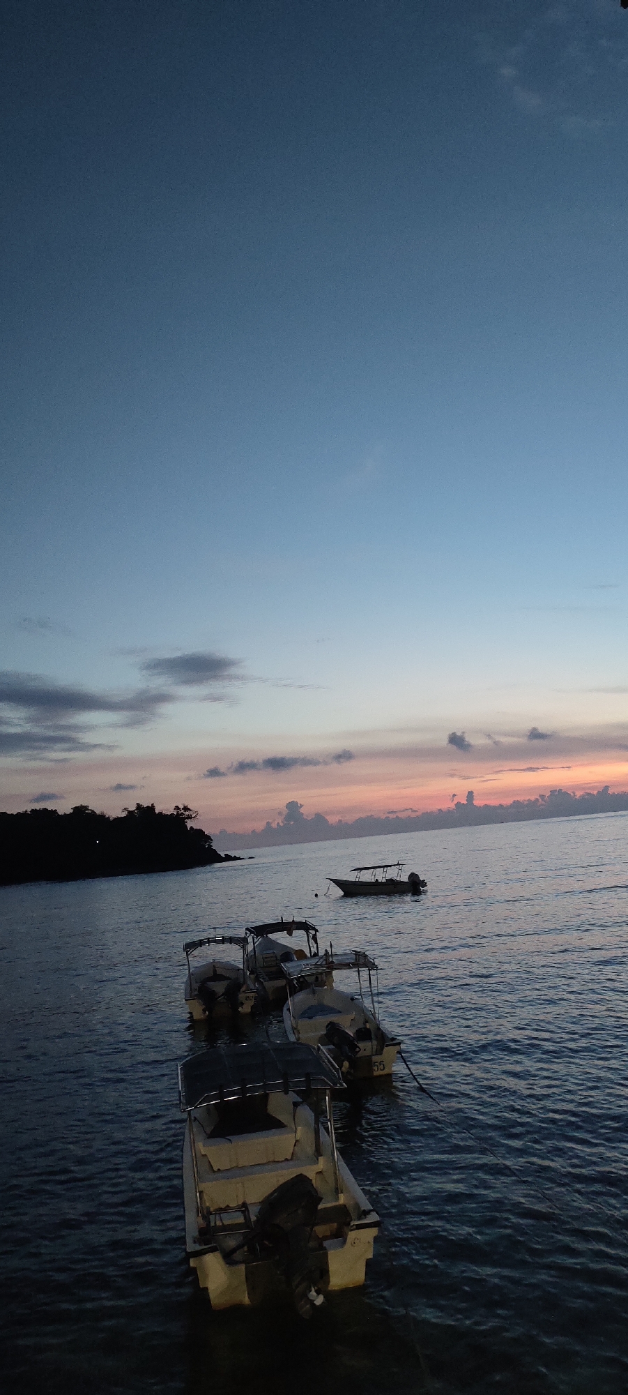 At dusk, several motorboats are moored on the calm, dark water near a treeline that extends into the ocean, likely on the coast of Sabang, Indonesia. The sky above is a gradient of deep blue near the top, fading to a soft pink and orange near the horizon, suggesting the sun has recently set. Small, scattered clouds are visible in the twilight sky. The boats appear to be of the same model, with white hulls and outboard motors. One boat is tied to another with a dark rope. Another boat is further out in the water, closer to the horizon. The scene is serene and peaceful, capturing the quiet beauty of a tropical evening.