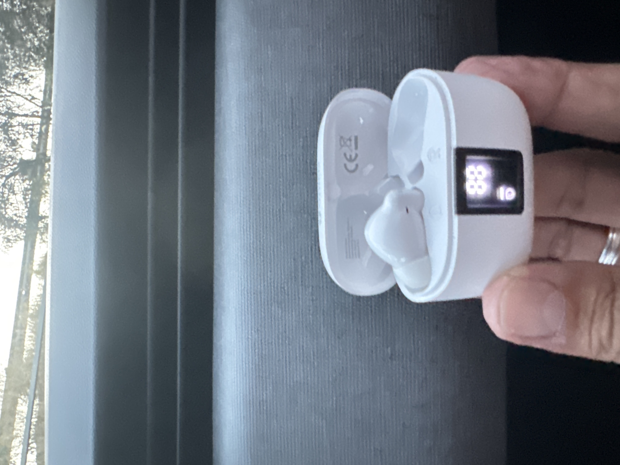 A close-up, low-angle, indoor shot shows a person's thumb and index finger holding a white wireless earbud charging case. The case is open, revealing a single white earbud inside. The case has a small digital display on the front showing "88" with a battery icon below it. The background is dark and textured, possibly a car's dashboard. The upper portion of the image shows a blurry outdoor scene with trees and sky, suggesting the photograph was taken inside a vehicle. No specific location in Murcia, Spain is identifiable.