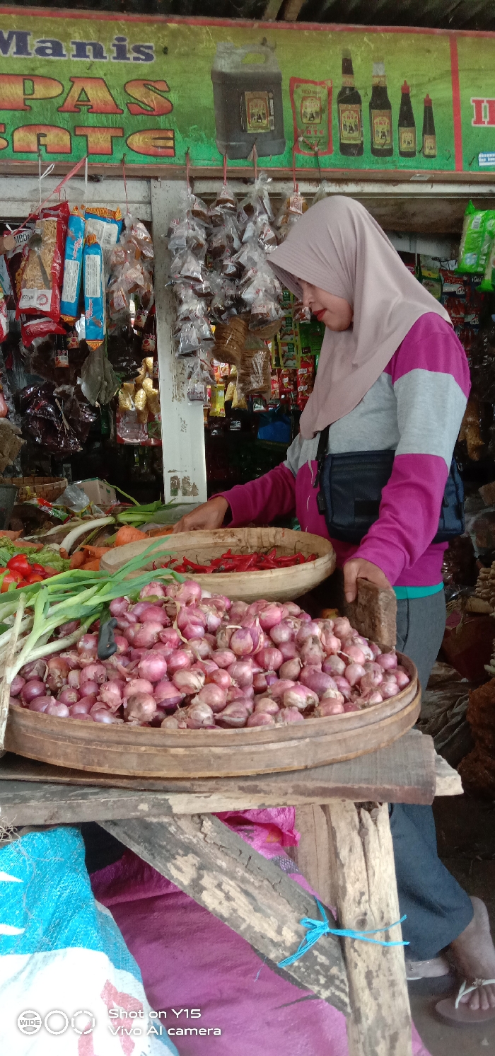 The image depicts an outdoor market stall in Maron, Indonesia, displaying fresh produce and packaged goods.
**People:**
One female individual is present, positioned behind a display of vegetables. She wears a light beige or pinkish-tan hijab, a long-sleeved shirt with a grey upper portion and magenta lower portion, and dark grey or black pants. A dark, rectangular fanny pack or crossbody bag is worn across her midsection. Her hands are visible, with her right hand near a basket of chili peppers and her left hand near a basket of shallots, suggesting interaction with the produce.
**Subjects/Objects:**
In the foreground, two circular, shallow woven baskets are stacked or placed closely on a rustic wooden stand. The lower, larger basket is filled with numerous small, red-skinned shallots. The upper, smaller basket contains a quantity of red chili peppers. To the immediate left of these baskets, various other fresh vegetables are visible, including green onions or scallions, carrots, and red bell peppers or tomatoes. The wooden stand appears to be constructed from rough, untreated planks.
**Background:**
Above the produce display, a green banner or sign with yellow/orange text is partially visible, reading "Manis" and "PAS SATE" or similar. The banner features illustrations of a large, dark container (possibly for soy sauce or cooking oil) and several bottles of soy sauce of varying sizes. Below the banner, numerous small items, individually packaged in clear plastic bags, hang from strings, likely snacks or condiments. More packaged goods are visible on shelves behind these hanging items. The stall's structure appears to be open or semi-enclosed. In the lower left foreground, a blue and white patterned woven material and a pink fabric are visible, possibly coverings or storage items.
