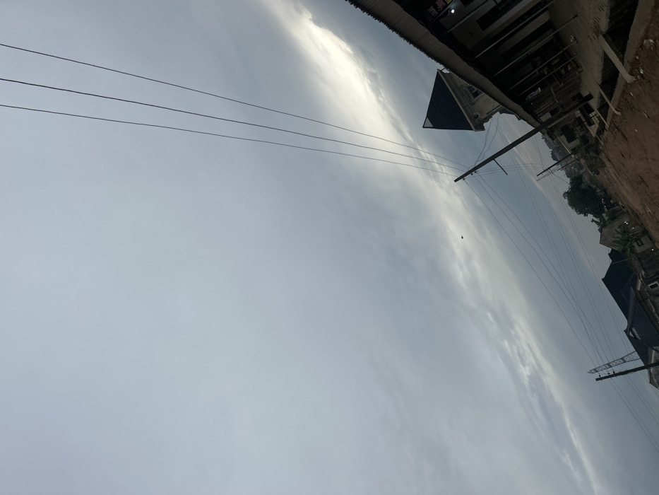 Overcast sky with a faint horizon line dividing the lower gray cloud cover from the upper lighter clouds. Power lines stretch across the sky. A building with a dark triangular roof is partially visible. Utility poles rise vertically. Residential buildings and trees line the ground in the background. A dirt path is visible at the upper right corner.