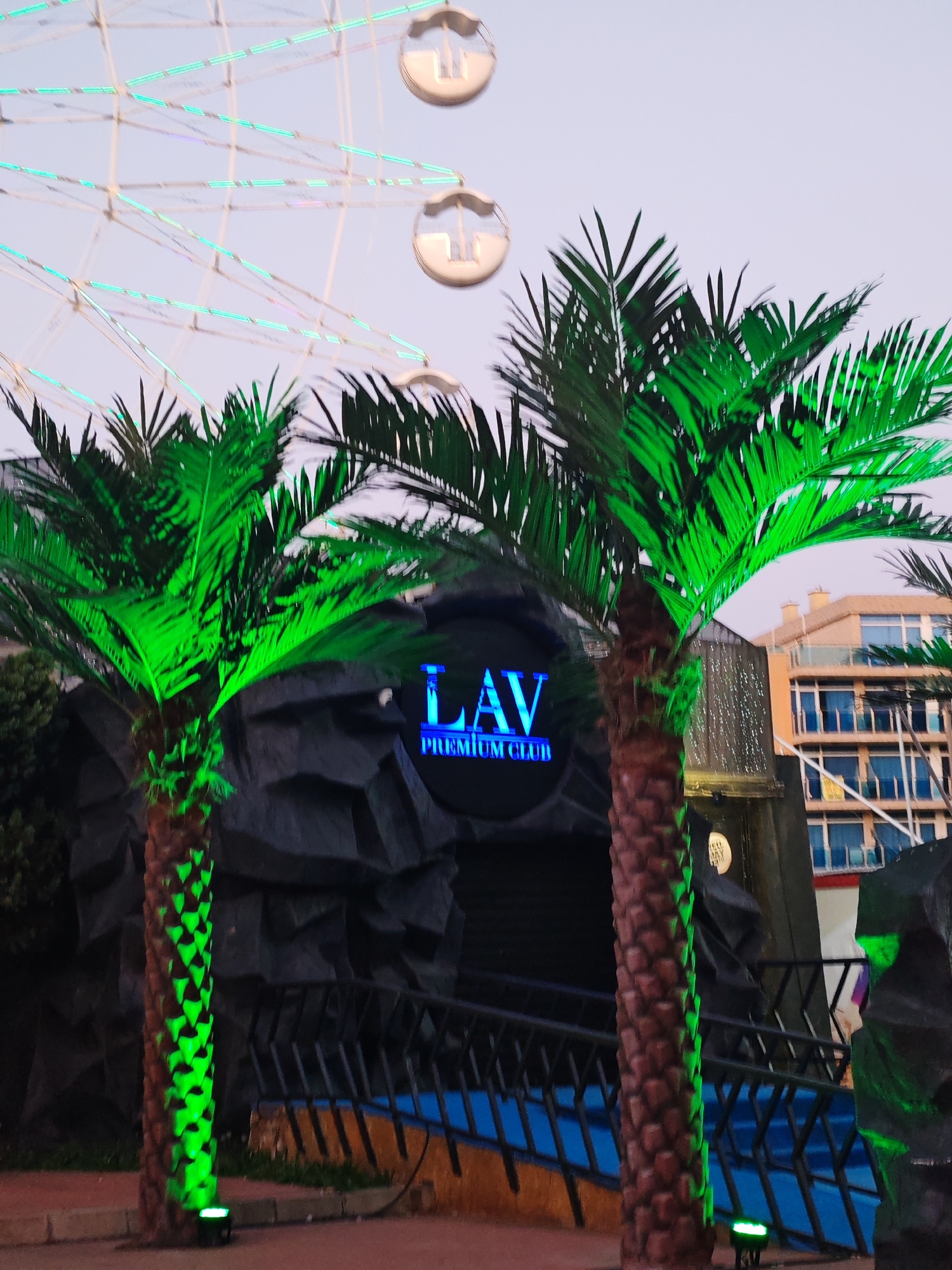 The image depicts the entrance to "LAV Premium Club" in Nesebar, Bulgaria. The entrance is framed by two illuminated artificial palm trees. Behind the entrance is a dark, rocky structure. In the background, a Ferris wheel with illuminated passenger cars can be seen. Also visible is a multi-story building with balconies and windows. The foreground includes a blue-painted railing. The time is dusk or evening, with soft, even lighting.