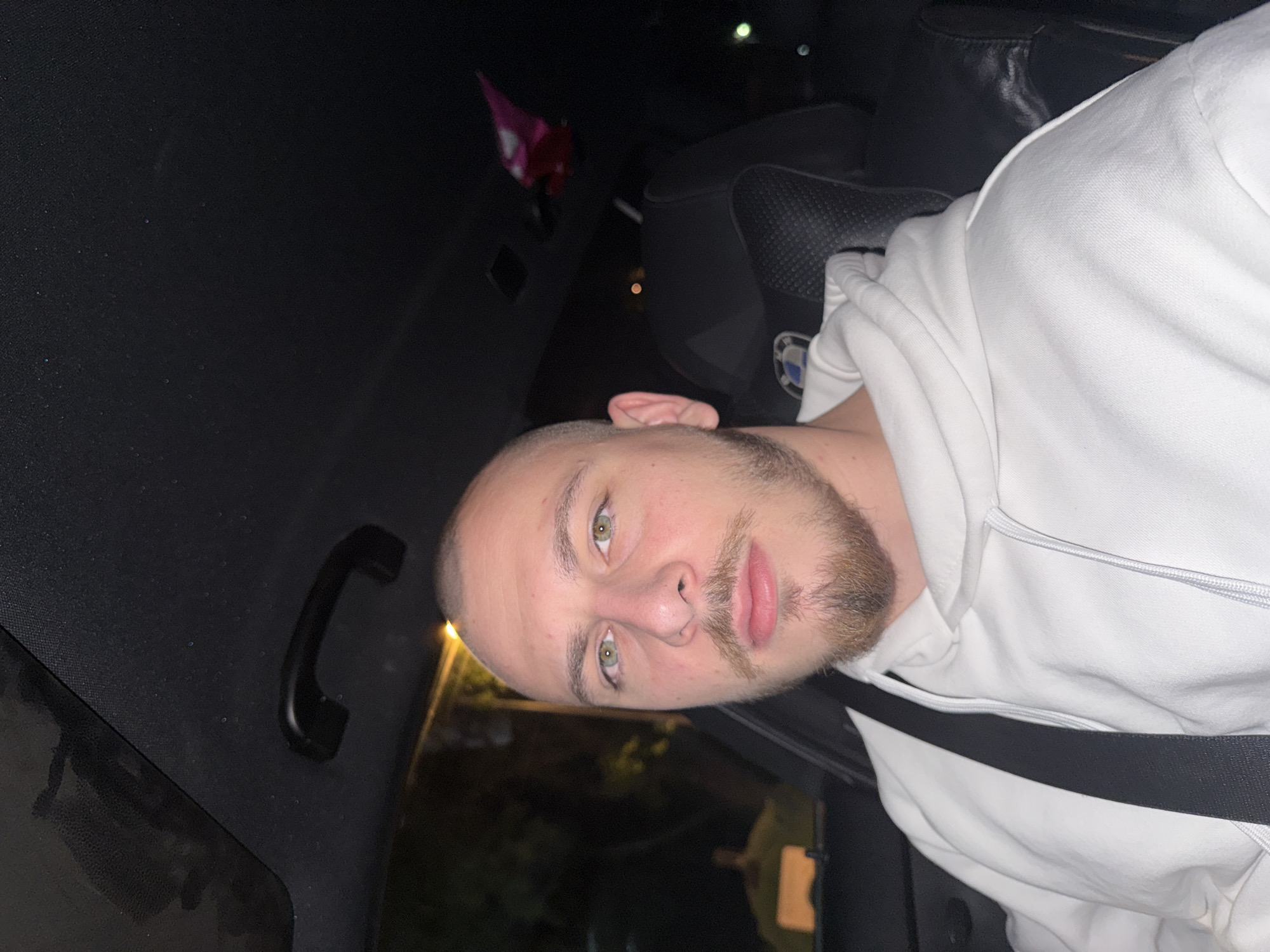 A man is in the driver's seat of a car, wearing a white hoodie and a seatbelt. The interior of the car is dark. Outside, there is some illumination, possibly from streetlights or buildings. The location is the city of Stara Zagora, Bulgaria. The image is taken from the passenger seat. The car is a BMW.
