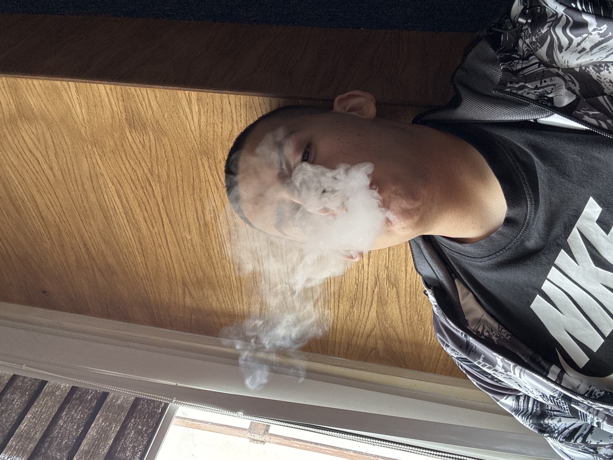 The image, rotated 90 degrees clockwise, shows a man indoors, exhaling a large cloud of white smoke or vapor. He is depicted from the head to approximately his chest, with a neutral expression as he releases the smoke. He has short, dark hair.
The man is wearing a black t-shirt with the word "NIKE" clearly visible in large white capital letters across the chest. He also has on a jacket with a distinctive black and white abstract or patterned design, worn open over the t-shirt.
The background consists primarily of a light-colored wooden surface, likely a wall or furniture, occupying the top and central areas of the frame. Below this, a window with horizontal blinds can be seen, indicating an indoor setting. The blinds appear to have a striped pattern. The lighting suggests natural light entering from the window. No specific visual cues for Plovdiv, Bulgaria are present within the image.