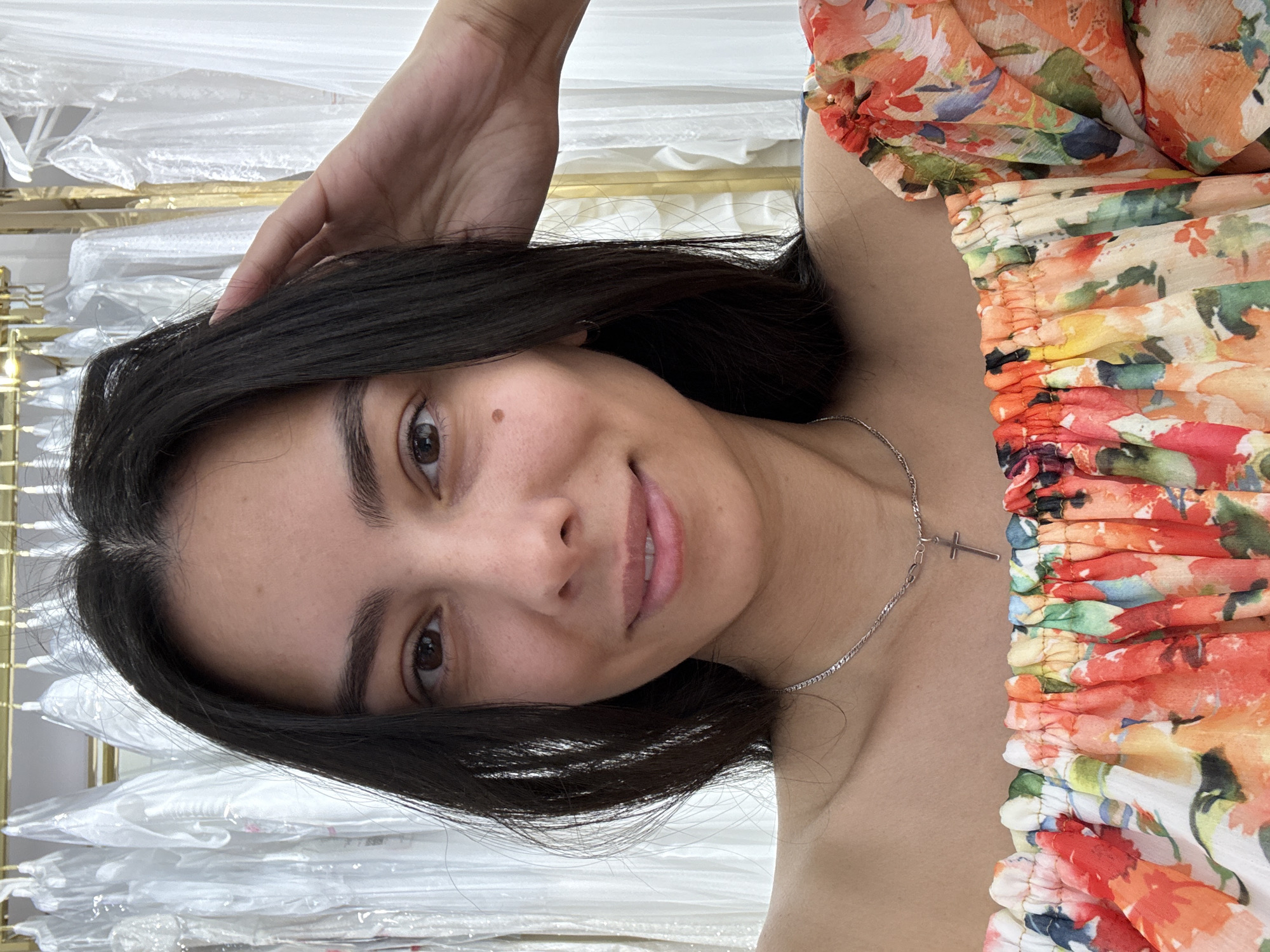 A woman with dark hair and a floral off-the-shoulder top is taking a selfie indoors. She is slightly tilted to the side with her hand in her hair. Behind her are rows of what appear to be white dresses or garments, possibly in a store. A silver necklace with a cross pendant is visible around her neck. There is no evidence to support this image being taken in the city of Chaikovsky, Russia.