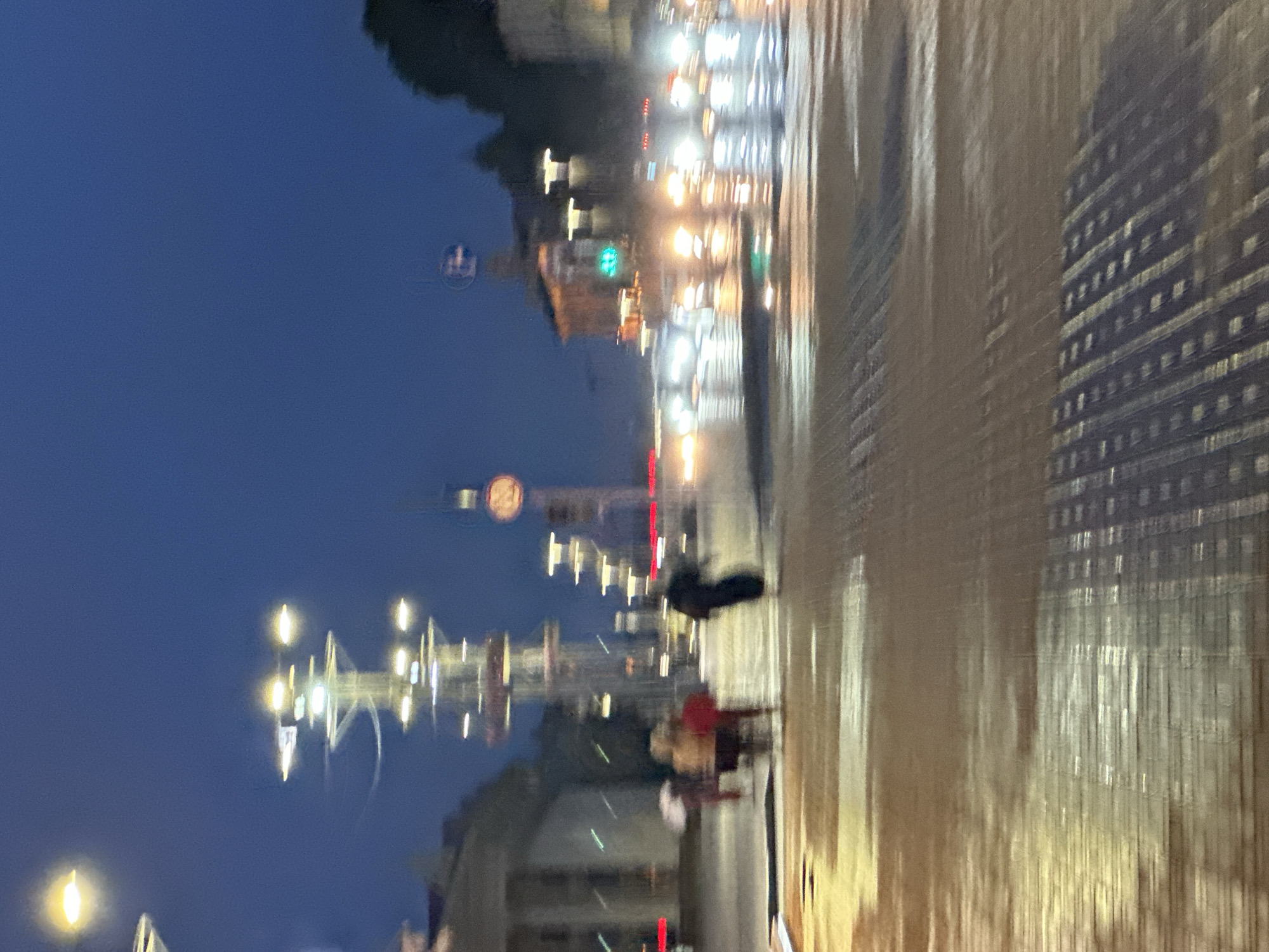 The image depicts a blurred nighttime scene of a city street in Minsk, Belarus, likely near the Triumph Arch or the Stella monument given the distinctive tall light poles and the general urban layout. The street appears wet, reflecting the city lights, suggesting it might have recently rained or is raining. Several blurry figures are visible, some crossing the street. One individual on the right is walking away from the camera, appearing to be wearing dark clothing. To the left, a few figures are grouped together, one holding a white umbrella. In the background, tall, multi-tiered light poles with multiple bulbs are prominent. The sky is a deep blue, indicating twilight or early night. The overall impression is one of a busy, yet somewhat desolate, urban environment at night.