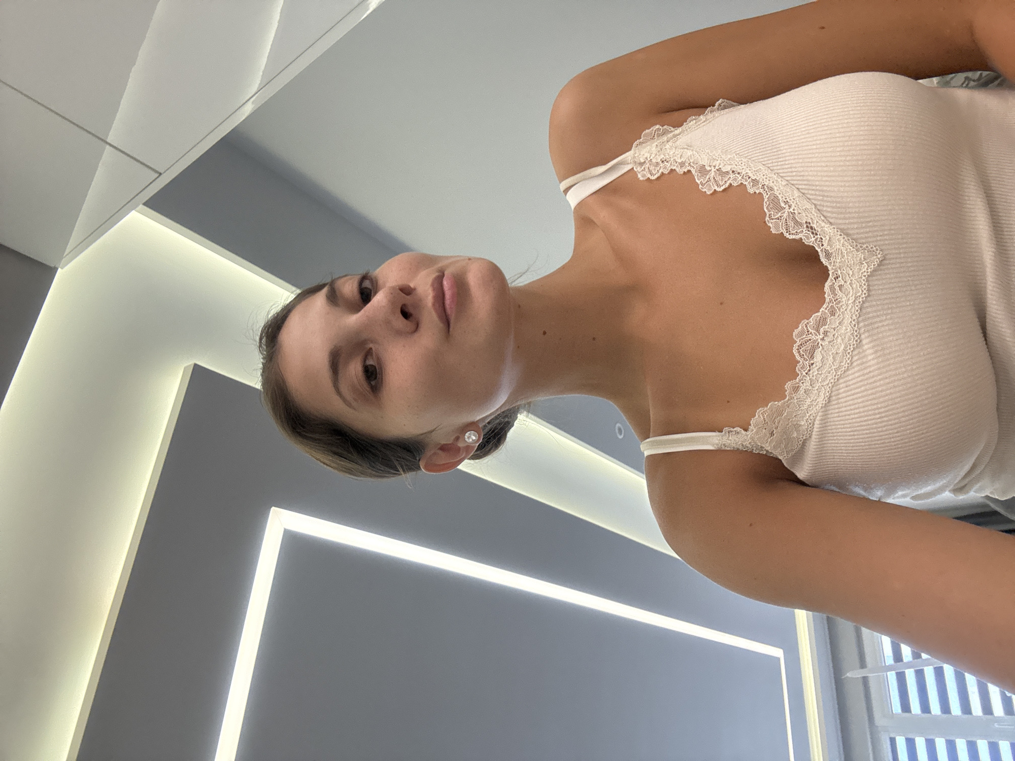 A low-angle indoor shot depicts a woman wearing a white tank top with lace trim. The background consists of a gray wall with recessed lighting. Outside, there is a glimpse of windows. The image appears to be taken from a mirror or reflective surface. There is no information available about this image that relates to Burgas, Bulgaria.