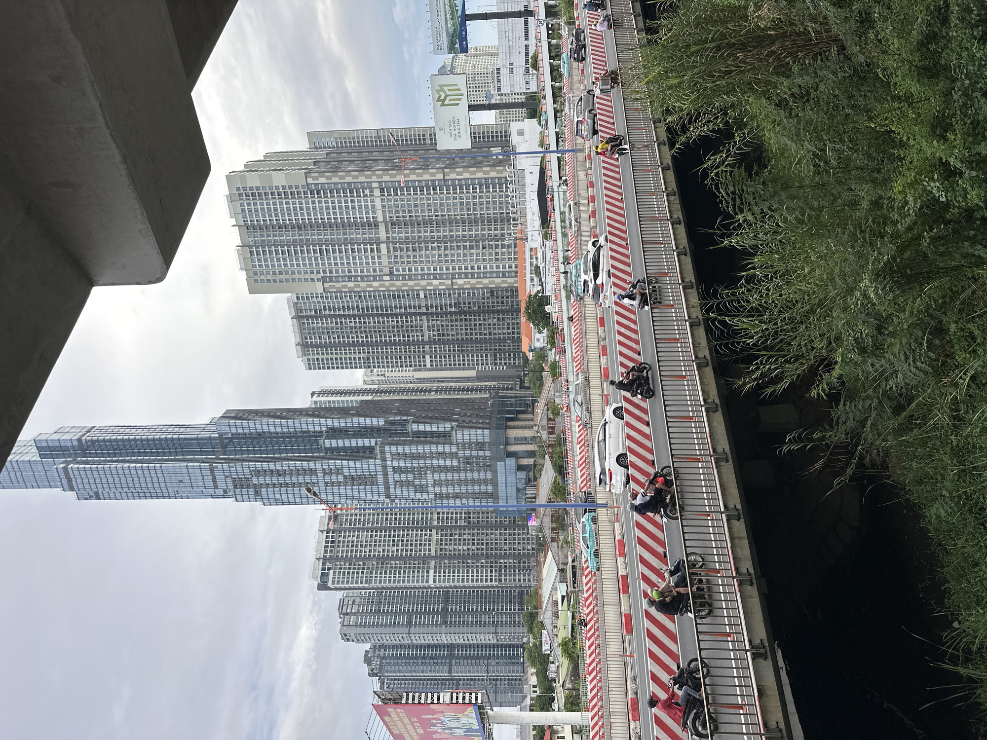 The image depicts a bridge in Thủ Đức, Vietnam, seen from an elevated perspective. The bridge is heavily trafficked with motorbikes and cars. The pavement is marked with alternating red and white diagonal stripes.
On one side of the bridge, tall, modern skyscrapers dominate the cityscape. A large billboard stands at the base of the buildings. On the other side of the bridge, dense greenery spills over the edge, obscuring the water below. The sky is overcast. A concrete structure is visible in the upper left corner of the image.