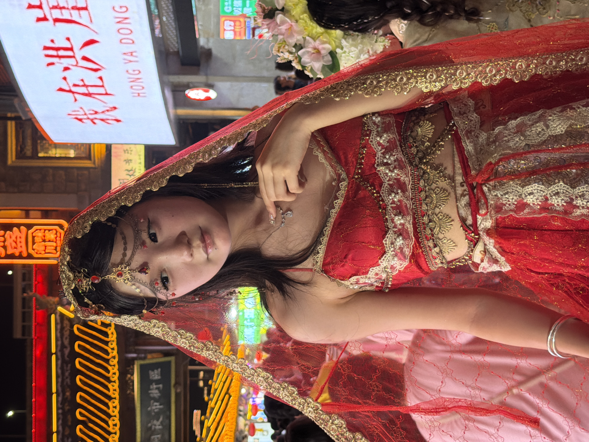A young woman is pictured at night, likely in Hongyadong, Yuzhong District, China, wearing a red and gold traditional outfit with a veil. She has intricate makeup on her eyes and a jeweled headdress. The background is blurred, showing neon lights and other people, suggesting a busy street or market. Her right hand is near her chest, and she is looking to her left. A bouquet of flowers is visible to the right of the frame. The lighting is artificial and vibrant, characteristic of an urban nighttime environment.