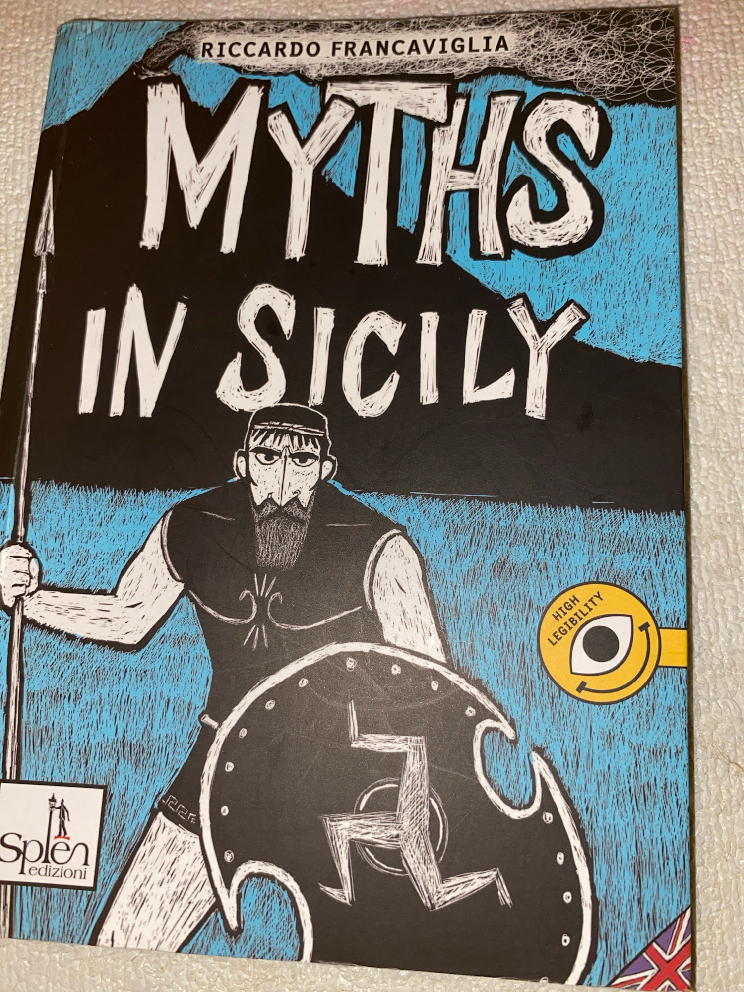 The image is a close-up of a book cover. The title, "MYTHS IN SICILY," is prominently displayed in large, white, distressed lettering against a black background. Above the title, in smaller black text on a white ribbon, is the author's name: "RICCARDO FRANCsp;AVIGLIA".
The artwork on the cover depicts a stylized drawing of a man holding a spear in his left hand and a shield in his right. The man has a beard, a headband, and wears a sleeveless tunic. The shield is large and circular, with a design that resembles a swastika, albeit rendered in a white, angular style. The background is a textured blue, possibly representing a sky or landscape.
In the bottom left corner of the cover, there is a logo for "Splen edizioni" which features a silhouette of a person holding a lamp. To the right, there is a circular yellow emblem with an eye symbol and the words "HIGH LEGIBILITY" around it. The bottom right corner shows a partial image of the Union Jack flag. The book cover is placed on a textured surface, possibly a carpet or fabric. There is no information in the image that indicates the location in the city of Barcellona Pozzo di Gotto, Italy.