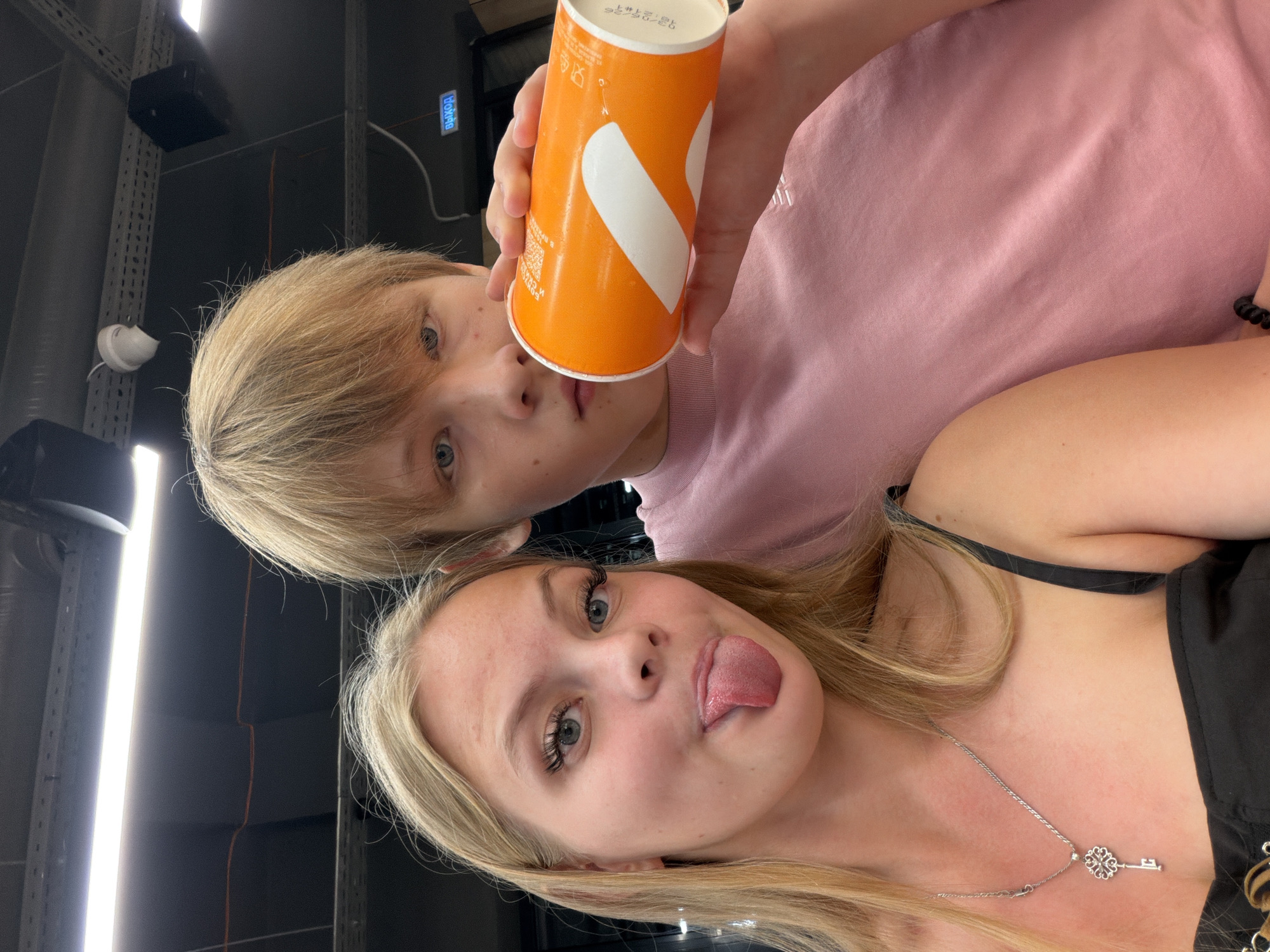 In Izhevsk, Russia, a young blonde woman with blue eyes and long eyelashes, sticking her tongue out playfully, poses for a selfie with a young man. She is wearing a black camisole and a silver necklace with a key-shaped pendant. The man, also with blond hair and blue eyes, holds an orange cup to his lips and wears a pink t-shirt. They are positioned in what appears to be an indoor public space with dark ceilings, fluorescent lighting, and exposed wiring, possibly a restaurant or retail establishment, indicated by a blue sign with the Cyrillic word "ВЫХОД" (EXIT) visible in the background.