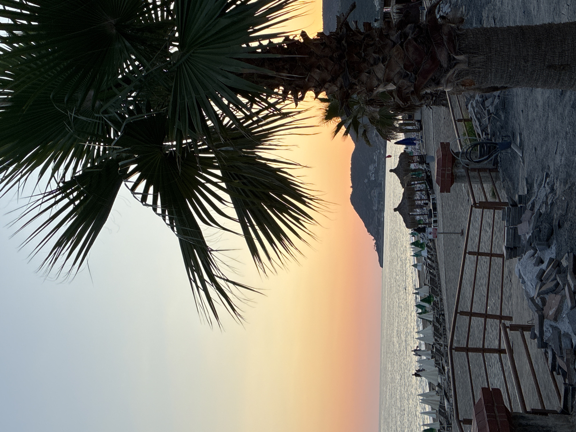 The image depicts a serene beach scene at dusk in Alanya, Turkey. In the foreground, a large palm tree trunk is visible on the right, its fronds extending upwards and obscuring parts of the sky. The ground is a mix of sand and construction debris, with a wooden railing and brick pillars suggesting ongoing or recent work.
Beyond the foreground, a sandy beach stretches out, lined with rows of lounge chairs and umbrellas. Some of the umbrellas are white, while others are green, indicating different sections or rentals. Several thatched-roof huts are also present on the beach, likely serving as shaded resting spots or small businesses. A few people are visible on the beach, scattered among the chairs and near the water's edge. They appear to be enjoying the evening.
The sea is calm, with gentle waves lapping at the shore. In the background, a chain of hills or mountains forms the coastline, silhouetted against the vibrant sunset sky. The sky transitions from a pale blue at the top to a warm gradient of orange and pink near the horizon, a typical display of Alanya's beautiful sunsets. The overall atmosphere is peaceful and relaxing, capturing the essence of a beach evening.