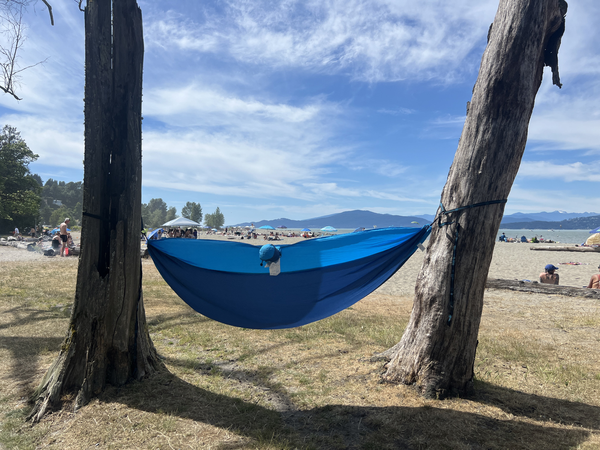 A bright blue hammock is strung between two weathered trees on a grassy area overlooking a sandy beach. The hammock is in the foreground, taking up a significant portion of the frame. In the background, the beach is dotted with people, beach umbrellas, and tents under a partly cloudy blue sky. Beyond the beach, a calm body of water is visible, with mountains in the far distance. The overall impression is a relaxed summer day at the beach.