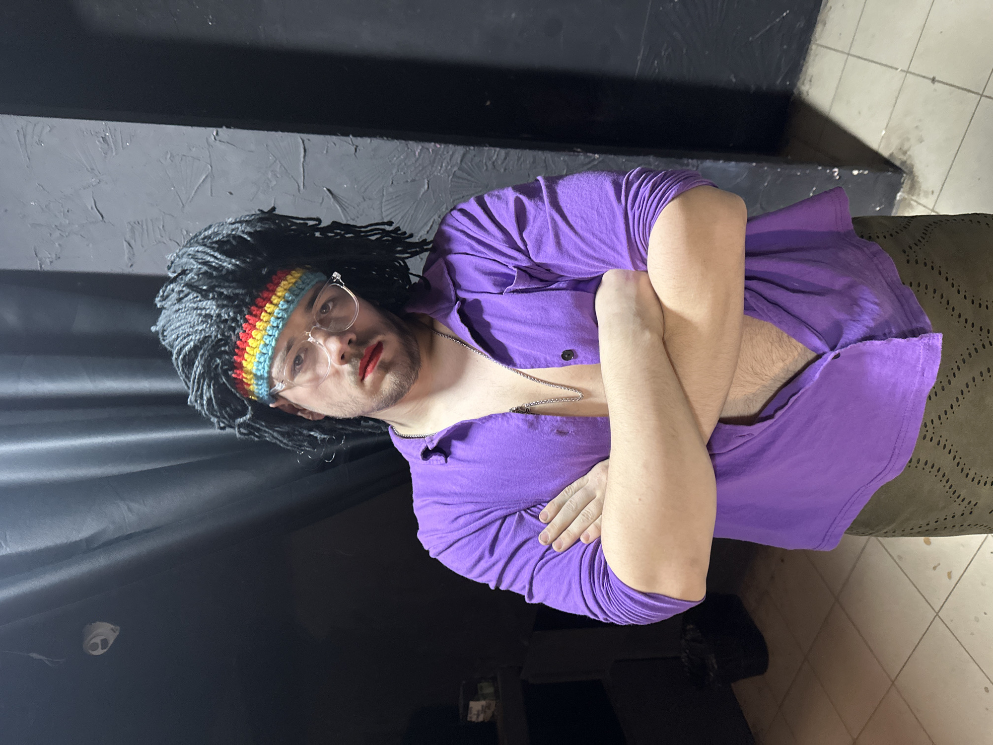 The image portrays a single individual, an adult male, standing indoors in the city of Saratov, Russia, facing the viewer directly. He wears a dark, possibly synthetic, wig styled with dreadlocks or braids, secured by a crocheted headband featuring horizontal stripes in red, yellow, light blue, and dark blue. Clear-framed eyeglasses are worn on his face, which also exhibits light facial hair on the chin and upper lip. Red lipstick is applied to his lips, with a noticeable smudge present on the upper lip. He is dressed in a long-sleeved, collared purple shirt, unbuttoned from the collar down, exposing a silver-colored chain necklace and a portion of his chest and abdomen. His arms are crossed in front of him. He wears dark green or brown trousers with a distinct patterned texture of small, regularly spaced perforations or raised dots.
The immediate background on the left features dark, vertical fabric drapes or curtains, with a small, light-colored, spherical object (possibly a sensor or camera) mounted high on the wall or ceiling near the top left corner. The background on the right consists of a dark-colored wall with an uneven, textured finish. The floor is tiled with light-colored, square tiles and dark grout lines, showing visible scuff marks and dirt accumulation. A dark waste receptacle is partially visible in the dim lighting of the far-left background. The scene is illuminated by an artificial light source, casting shadows behind the subject.