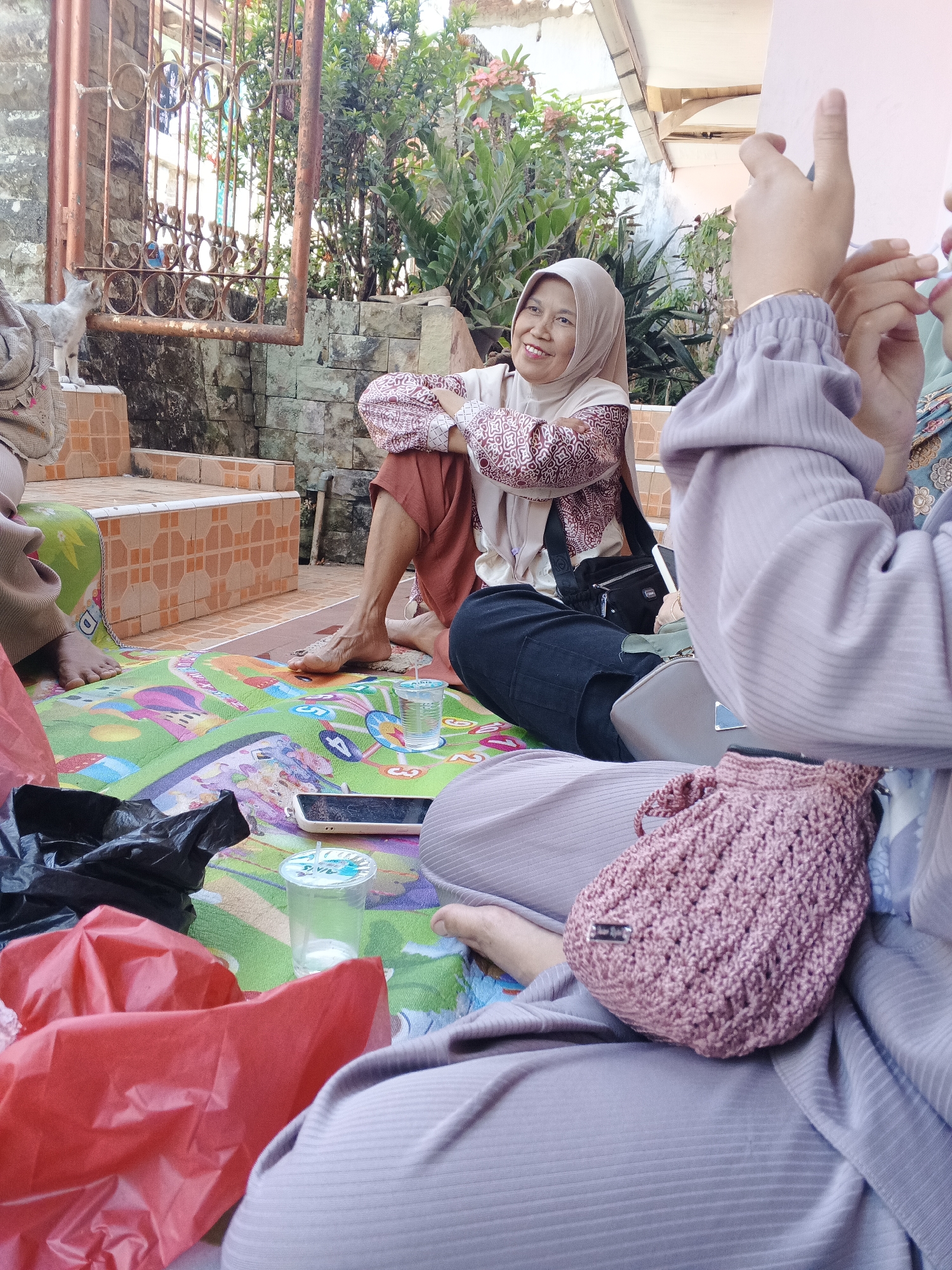 In Warungkiara, Indonesia, four individuals are gathered outdoors, seated on a colorful mat spread on the ground. The setting appears to be a residential area, with a stone wall and lush greenery visible in the background. One woman, wearing a light brown hijab and a patterned top, is seated with her arms crossed, smiling. She is positioned near a set of orange tiled steps leading up to a rustic metal gate. In the foreground, two other individuals are partially visible, their bodies covered by light purple clothing. A pink crocheted bag rests on the lap of one of them. Scattered on the mat are various items, including a transparent plastic cup with a blue liquid, a smartphone, a black plastic bag, and a red plastic bag. A small, light-colored cat stands on the edge of the tiled steps, looking towards the gate. The overall atmosphere suggests a casual gathering or a moment of rest outdoors.