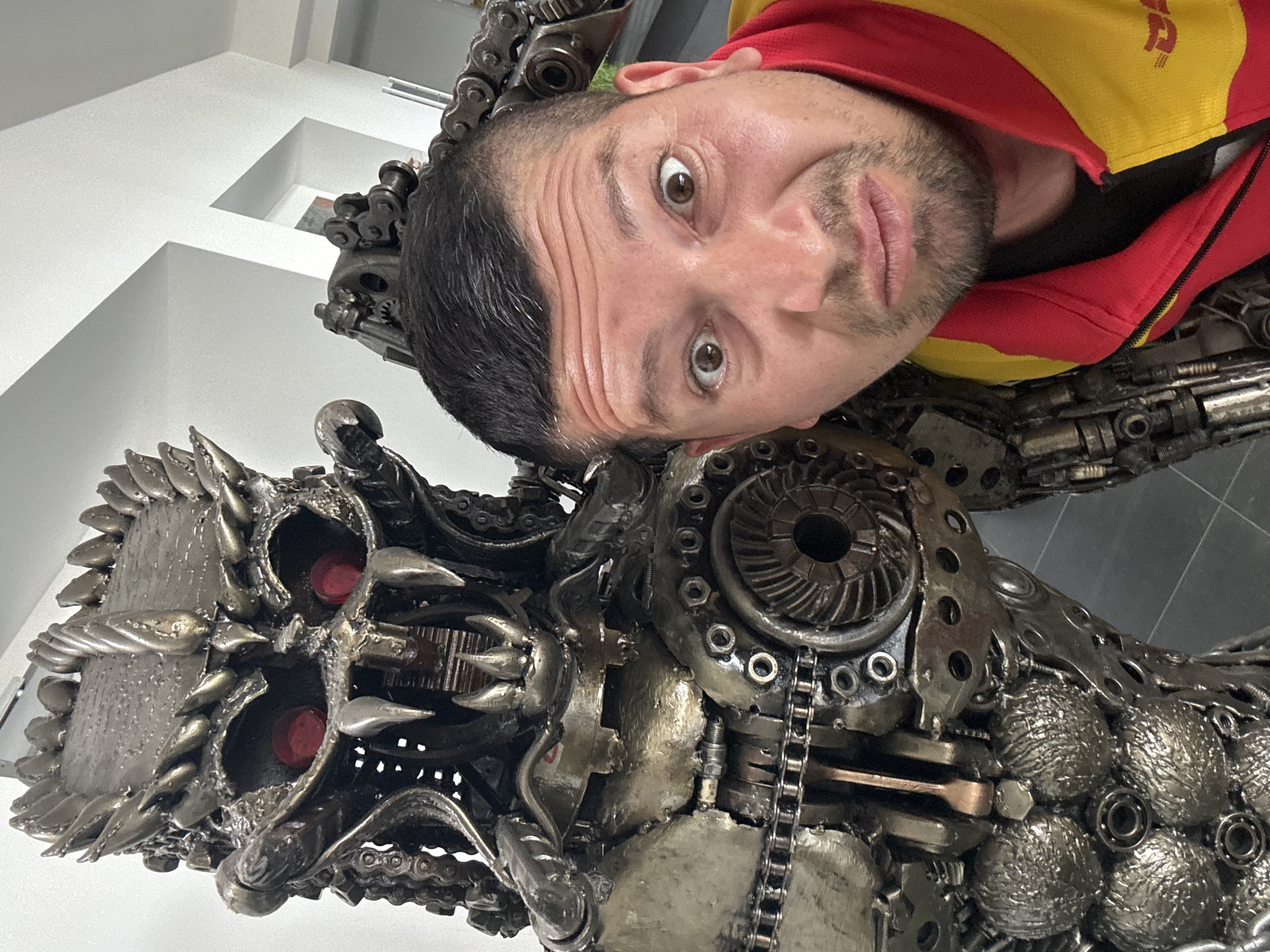 A man in a red and yellow zip-up jacket poses for a selfie with a metal sculpture in Bietigheim-Bissingen, Germany. The sculpture, made of scrap metal parts, has a menacing, skull-like appearance with red-painted eye sockets. The man looks directly at the camera with a slightly puckered expression. The indoor setting has white walls and a grid-like floor.