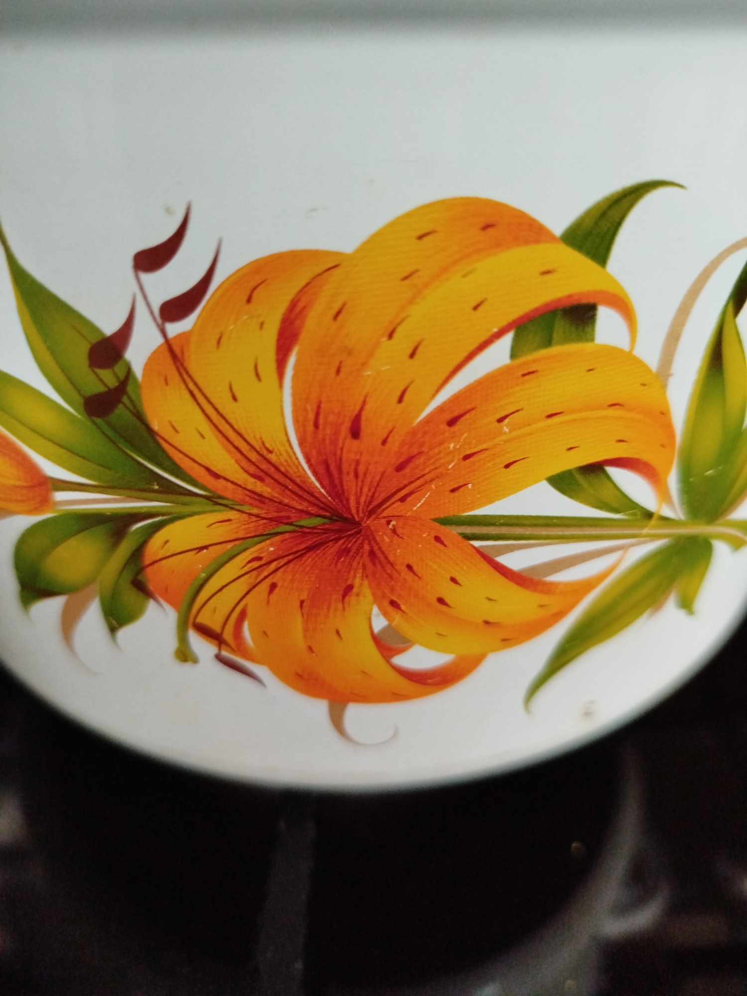 The image is a close-up shot of a decorative floral pattern on a white surface, likely a ceramic or enameled item. The pattern features a vibrant orange and yellow lily with intricate red speckles on its petals. Green leaves and thin brown stems complement the flower. The background is a soft white, with the bottom of the image transitioning to a dark, out-of-focus background. The lighting suggests an indoor setting, possibly a kitchen or dining area, given the glossy finish and the implied context of decorative household items. The specific location in Крывасельскі сельскі Савет, Belarus cannot be determined from the visual information provided.