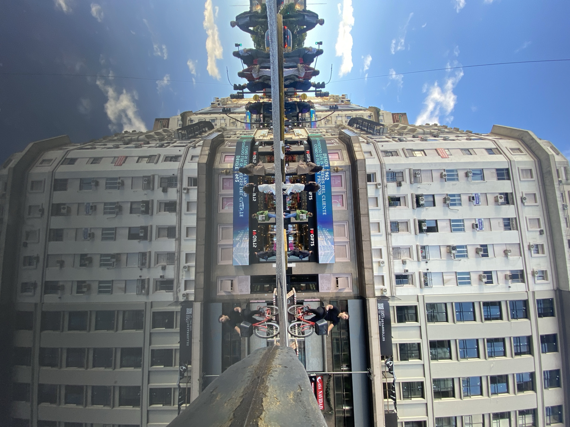 The image captures an architectural urban scene in Buenos Aires, Argentina, dominated by a large, light-colored building under a clear blue sky with scattered white clouds. The photograph employs a striking reflection, likely from a large, dark, vertical reflective surface (possibly a polished building facade or monument) in the foreground.
The right side of the image displays the actual multi-story building, characterized by numerous windows and external air conditioning units. Its structured facade features ledges and architectural details. The left side is a direct reflection of this same building and the sky. The central vertical strip, however, presents a complex reflection of the street scene opposite the viewer, including people, street infrastructure, and commercial banners, all appearing inverted and symmetrical along a central axis.
Prominently featured in this central reflection are several people, including one person lying horizontally with a red bicycle, creating a surreal visual effect. Street poles, traffic lights, and other urban elements are also visible in reflection.
Visible text on the building and banners, some appearing in reverse due to the reflection, includes "Urrutia" and "VENDE" (sells), "DESDE 1962 CUIDANDO LOS INTERESES DEL CLIENTE COMO SI FUERAN PROPIOS" (Since 1962, caring for the client's interests as if they were their own), "I❤GIFTS" or "I GIFTS," "B&R DESARROLLOS" (B&R Developments) with a phone number "Tel: 4890-8062," and "CASA NAVIDAD" (Christmas House). The bright daylight suggests it's midday or afternoon, and the weather is sunny. The overall composition creates an intriguing, almost kaleidoscopic view of an everyday city environment.